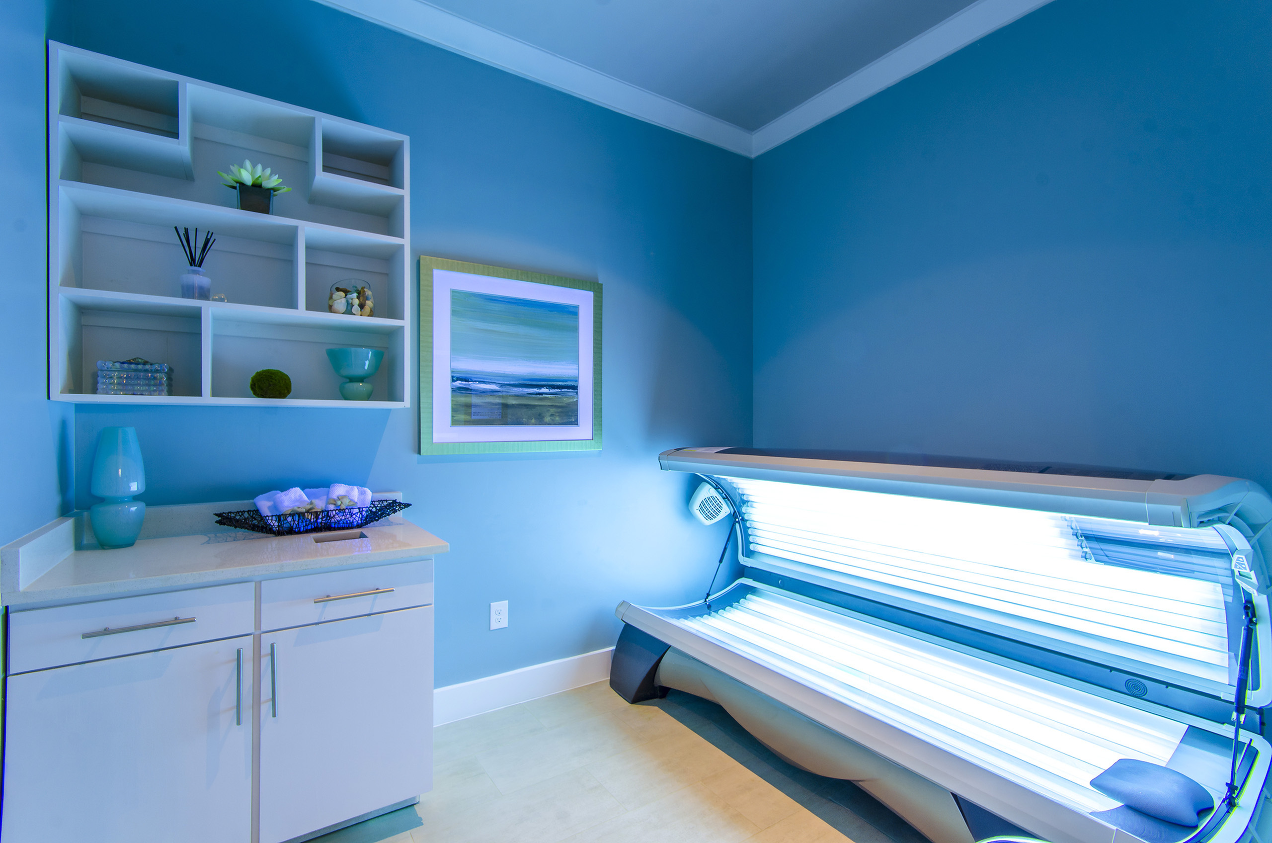 Modern tanning room with sleek sunbed and elegant blue decor, complemented by stylish shelving and artistic wall art.
