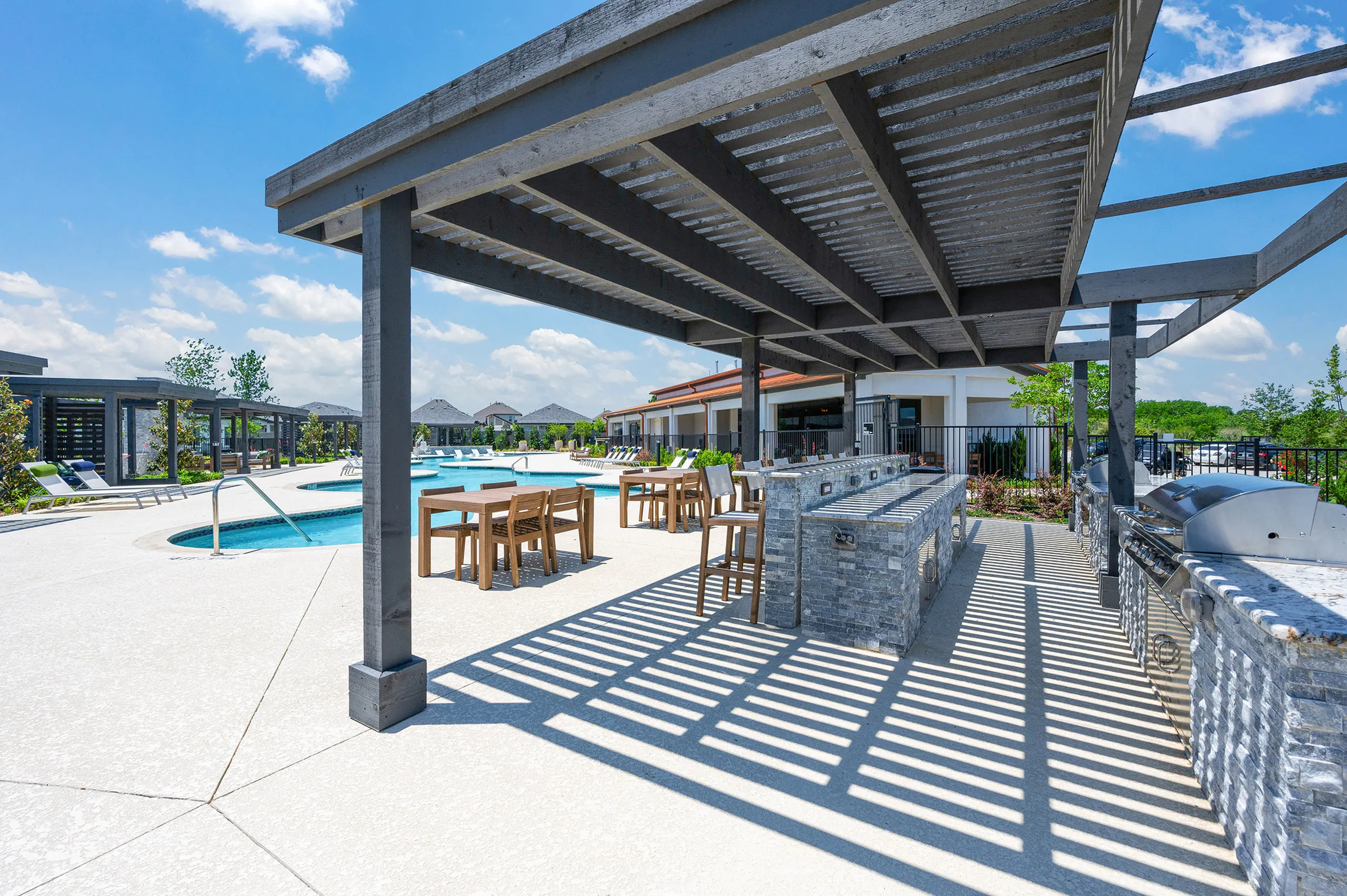 Luxurious outdoor pool area with shaded seating, barbecue grills, and lush greenery under a clear blue sky.
