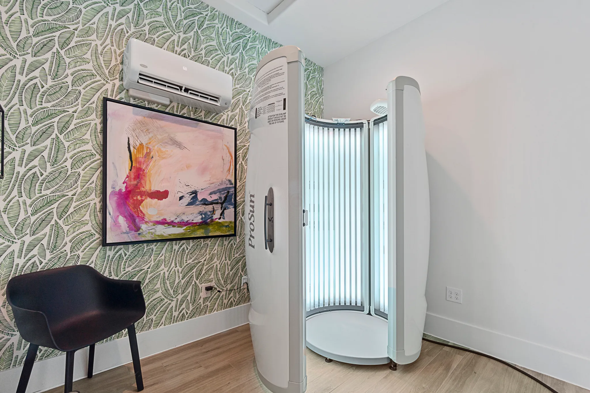 Stylish corner featuring a modern tanning booth with sleek white design, vibrant abstract artwork, and lush green leaf-patterned wallpaper.