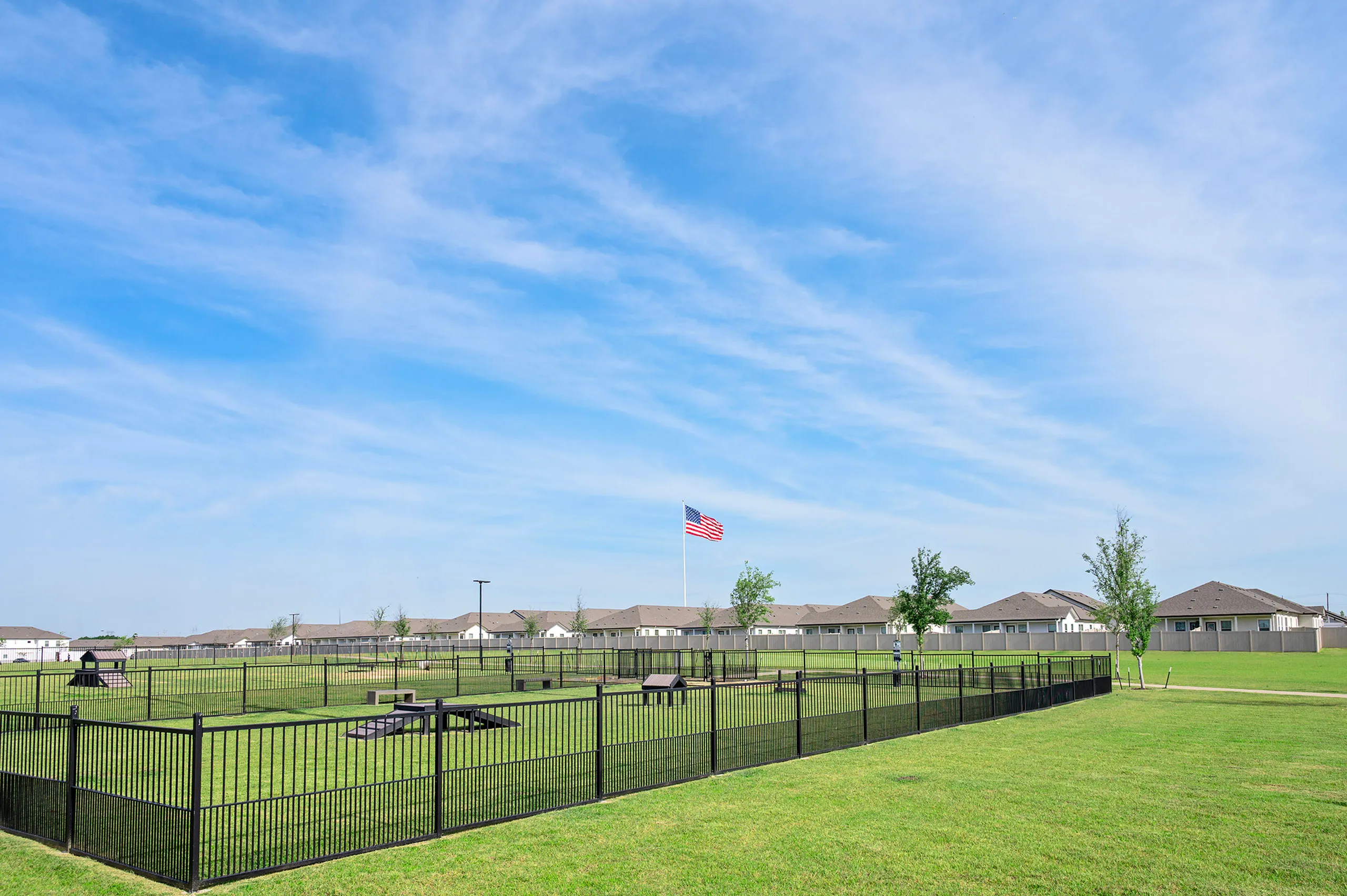 Expansive fenced green area with dog park features, perfect for outdoor activities, under a clear blue sky and American flag waving in the distance.