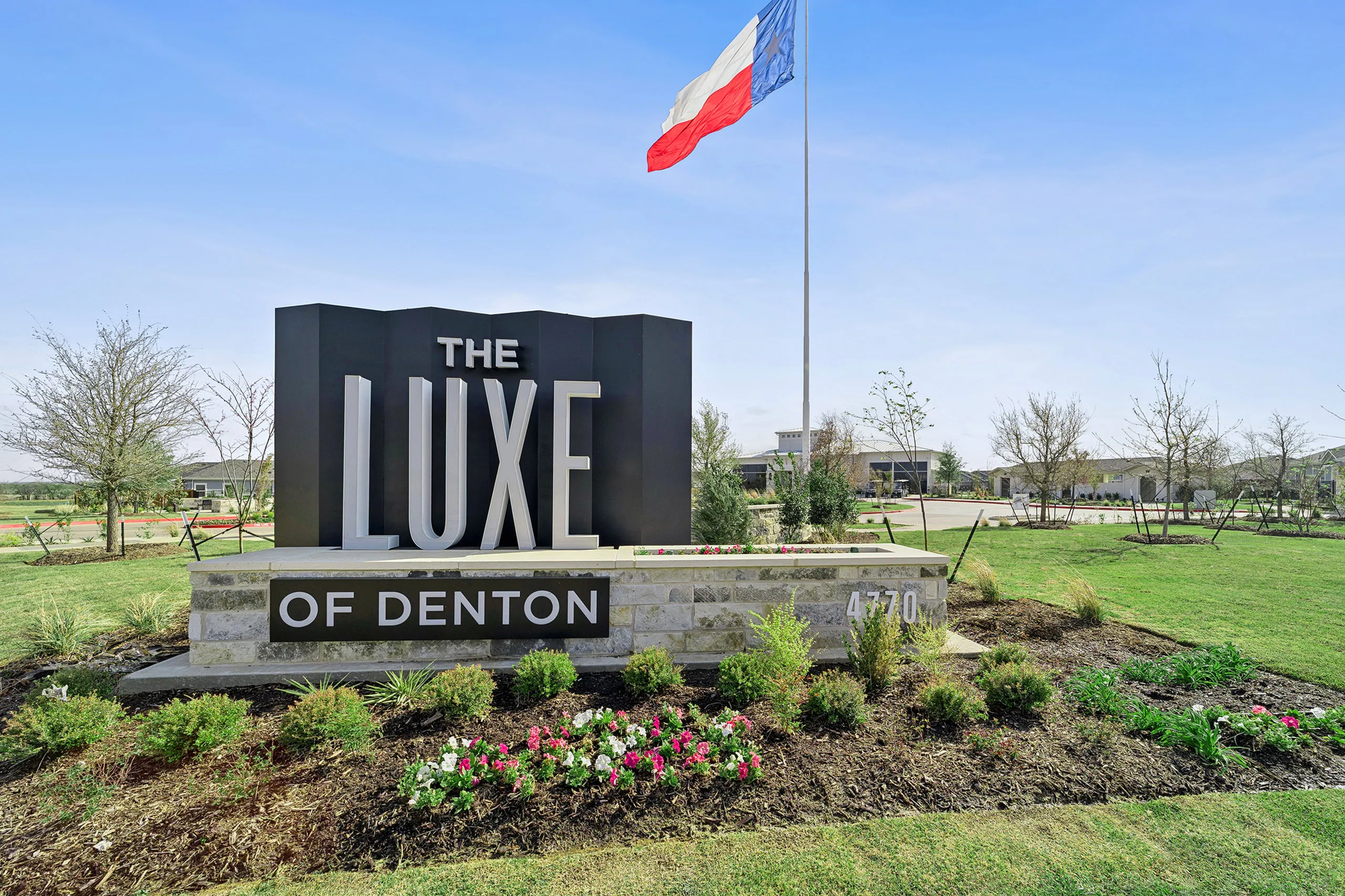 Inviting entrance to The Luxe of Denton, featuring elegant signage surrounded by lush green landscaping and vibrant flowers.