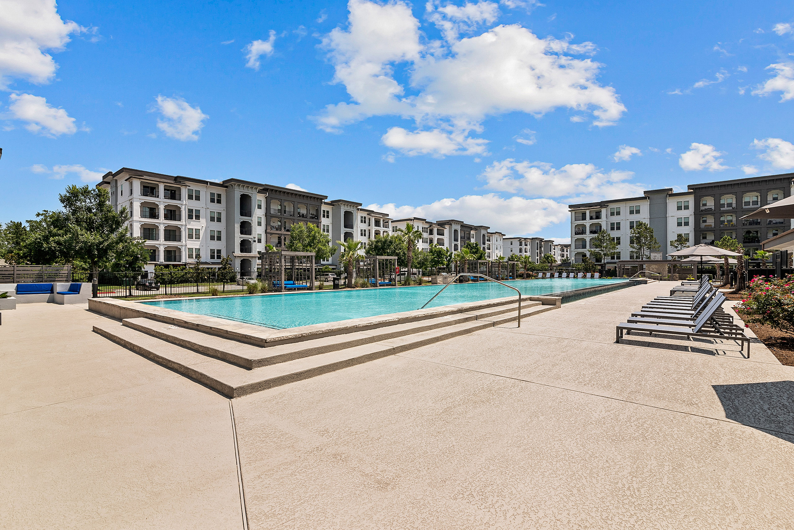 Luxurious apartment complex featuring a stunning outdoor pool, surrounded by elegant lounging chairs and lush greenery under a bright blue sky.