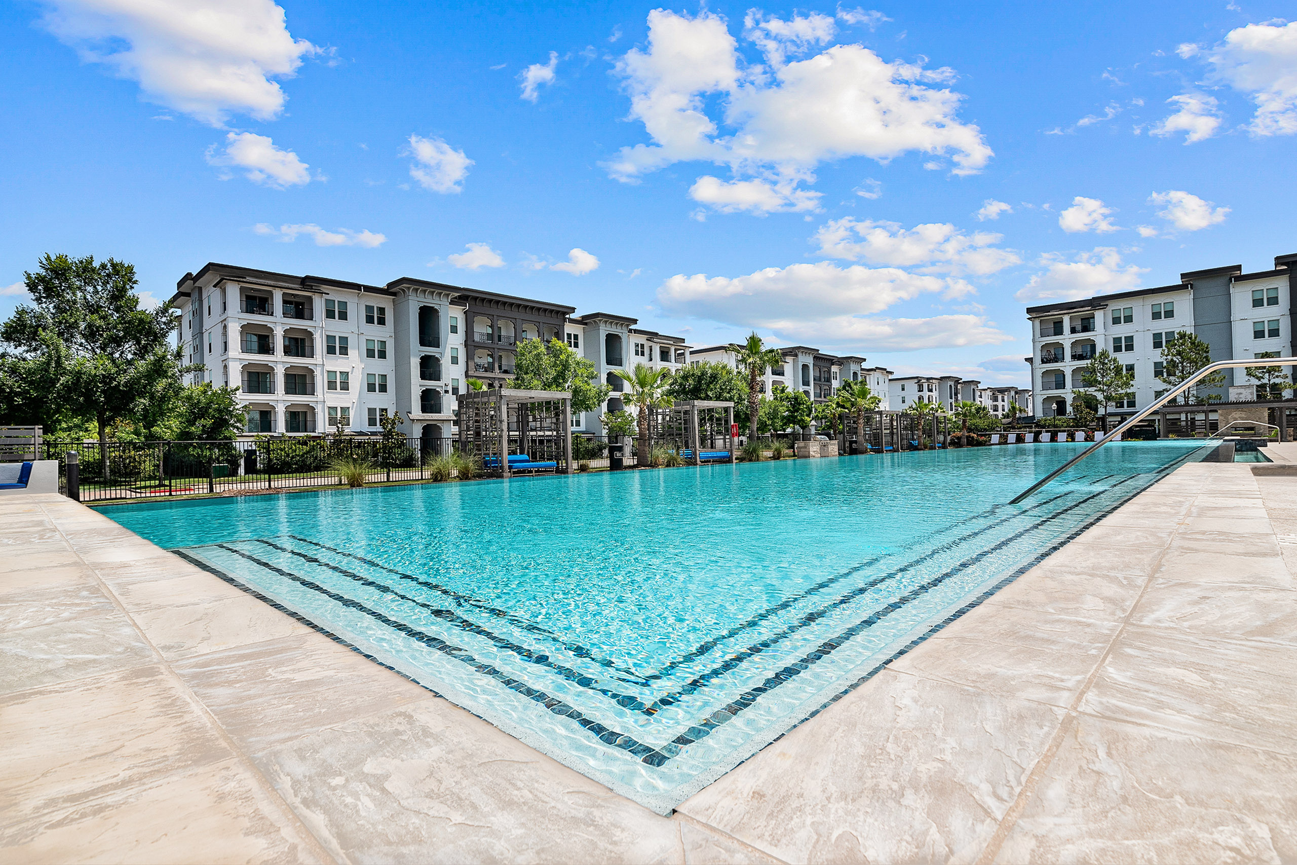 Luxurious apartment complex featuring a stunning resort-style pool with crystal-clear water, surrounded by elegant multi-story buildings and lush landscaping.