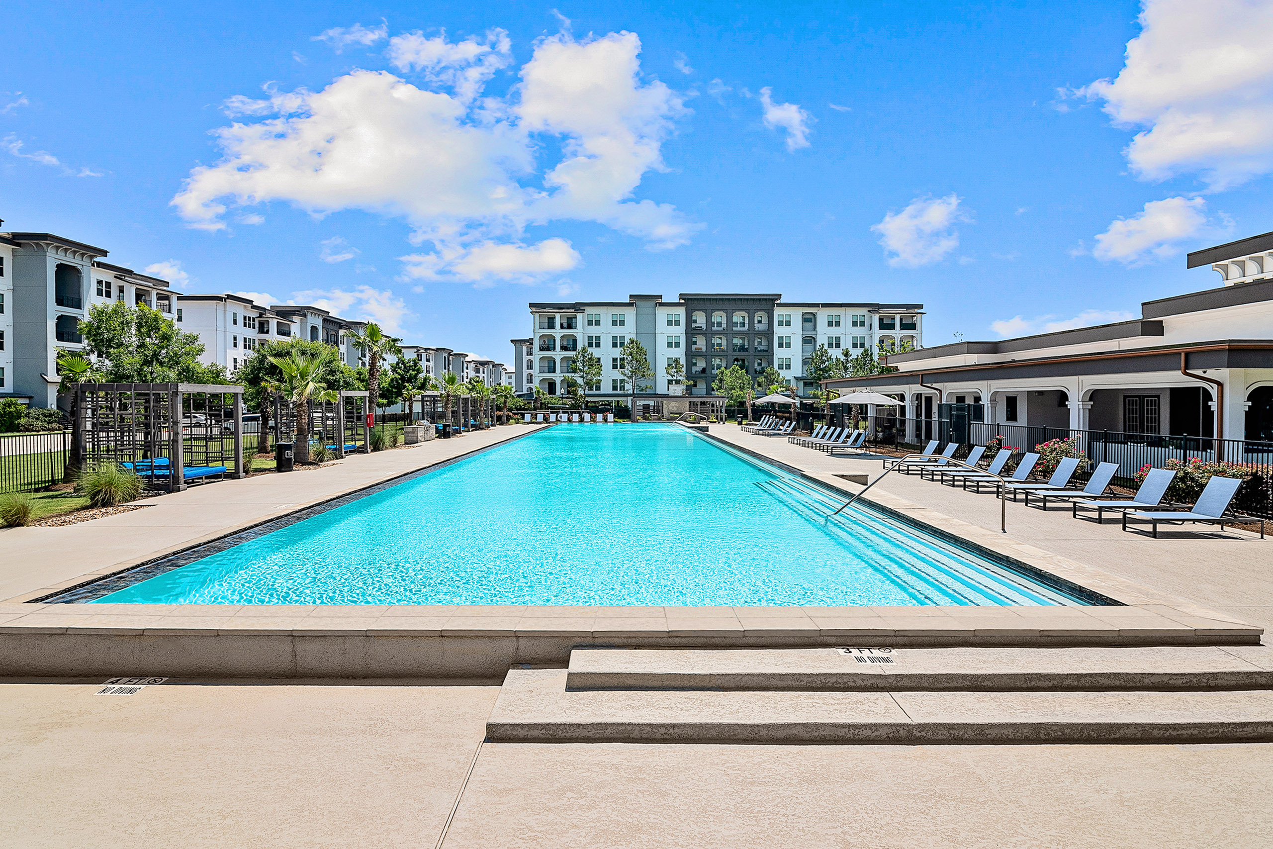 Luxurious apartment complex featuring a sparkling swimming pool with lounge chairs and modern architecture under a bright blue sky.