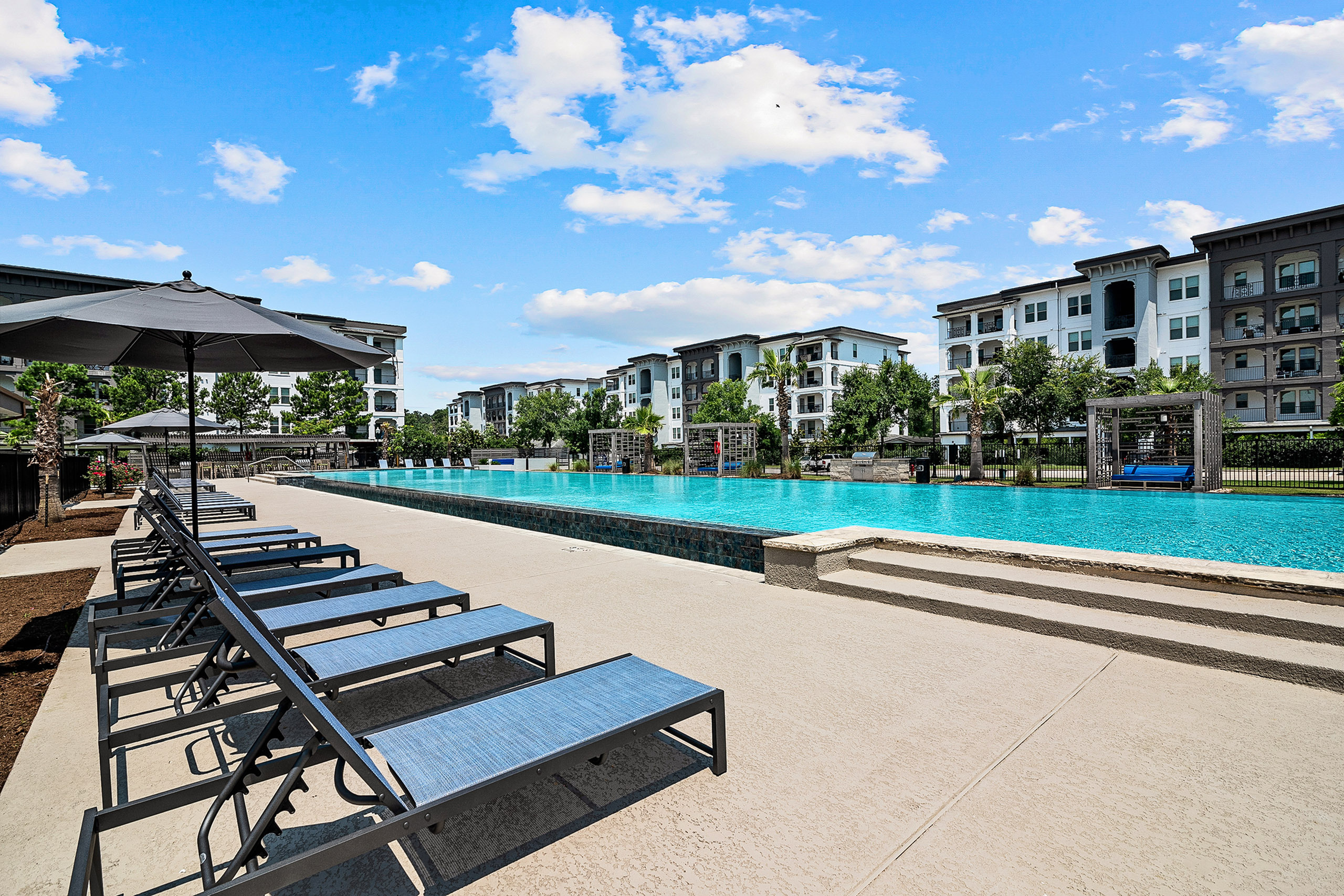 Luxurious apartment complex showcasing a stunning swimming pool, modern lounges, and vibrant outdoor space under a clear blue sky.