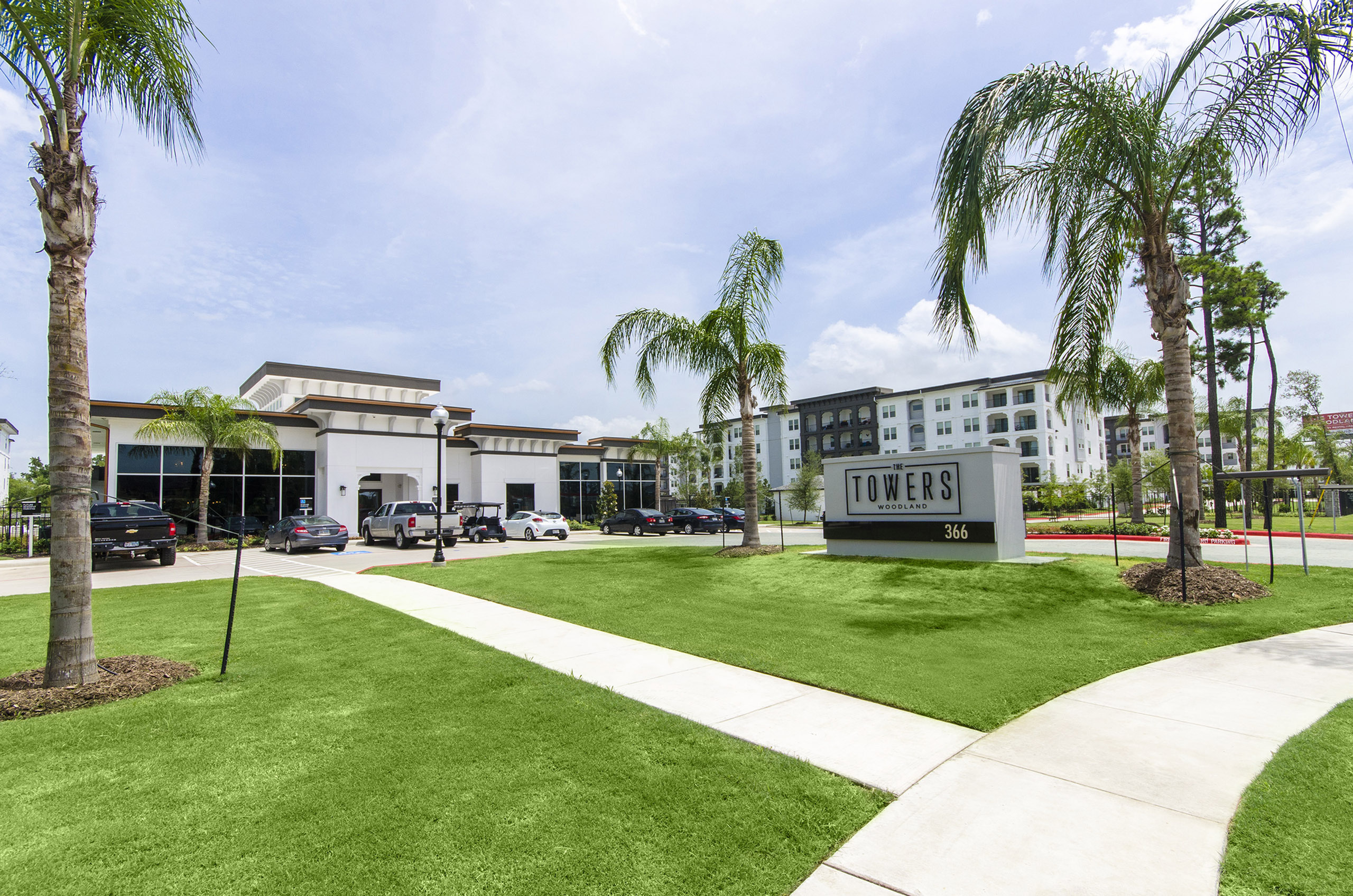 Inviting modern apartment complex entrance surrounded by lush green lawns and elegant palm trees, showcasing The Towers Woodland.