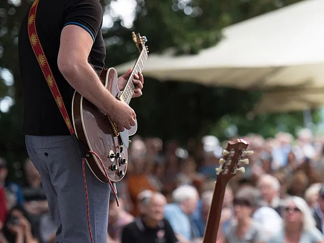 Musician playing electric guitar on stage, captivating a lively outdoor audience under sunny skies.