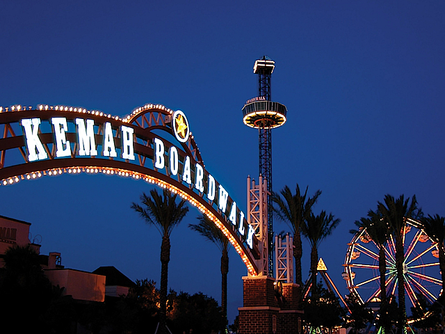 Vibrant night view of Kemah Boardwalk entrance, illuminated ferris wheel, and towering observation ride set against a deep blue sky.