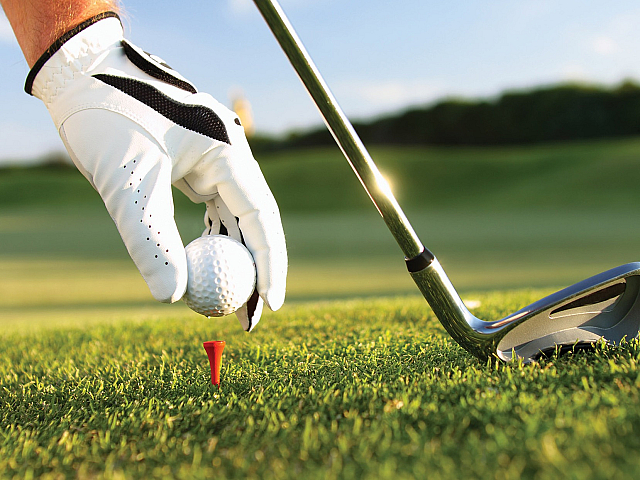 Close-up of a golfer's hand with a glove placing a golf ball on a tee with a club in the background, set against a lush green golf course under a bright sky.