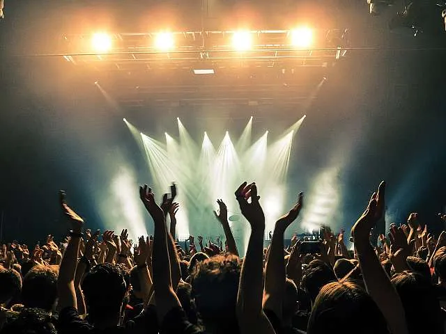 Energetic concert crowd with hands raised, illuminated by vibrant stage lights, creating a lively atmosphere.