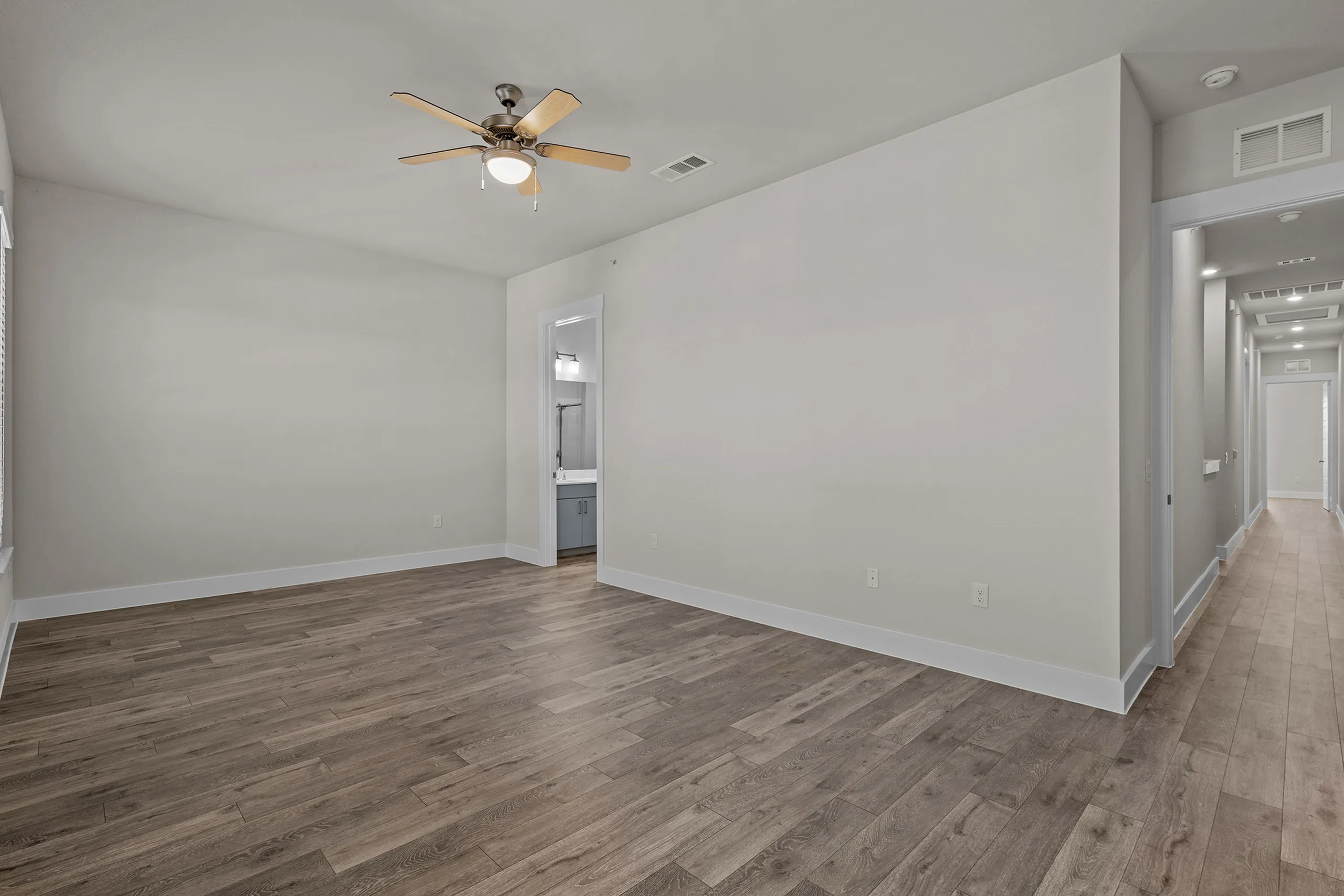 Spacious room with elegant wood flooring, modern ceiling fan, and bright hallway leading to a well-lit bathroom.