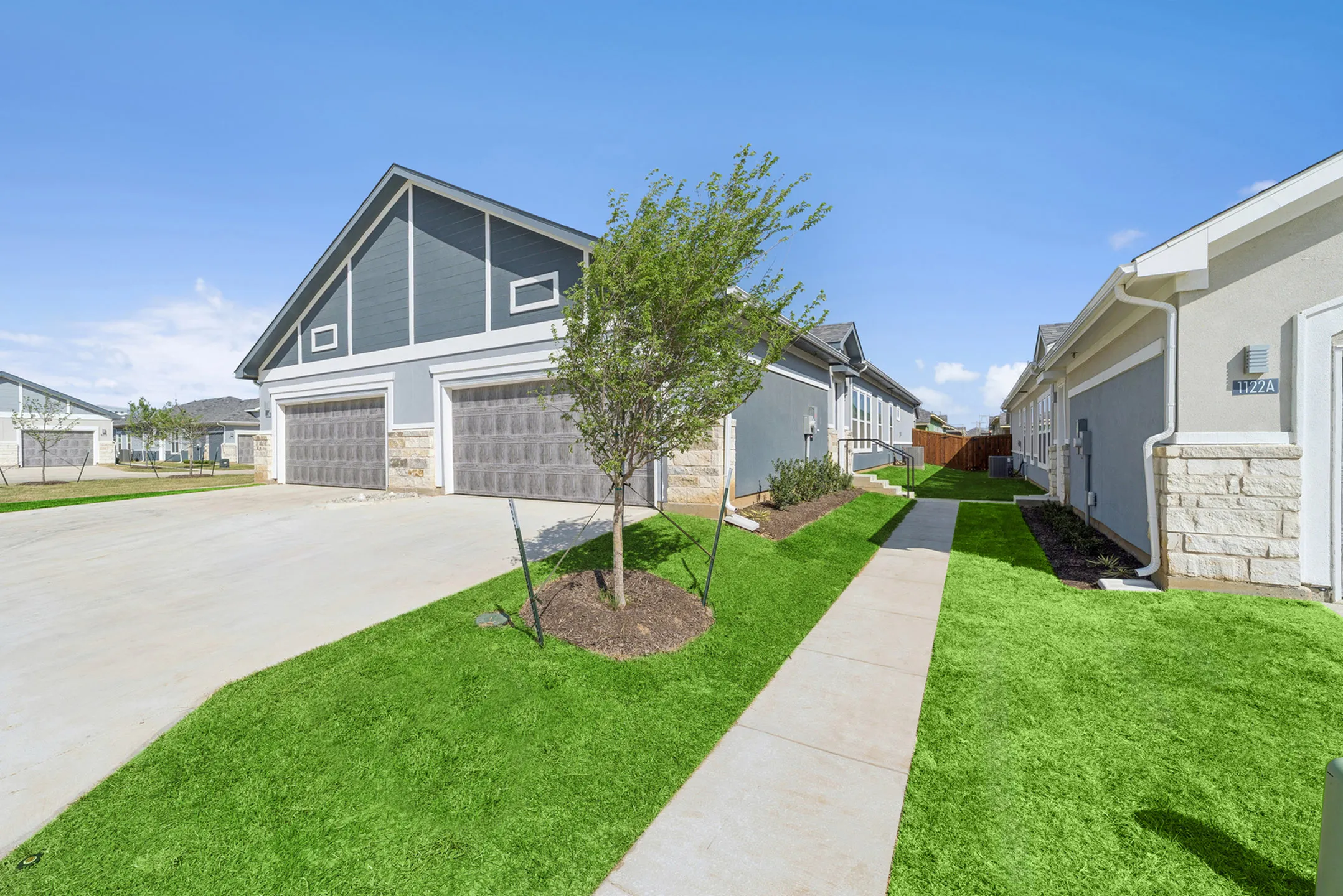 Contemporary apartment complex with modern gray exterior, double garage doors, and lush green landscaping under a clear blue sky.