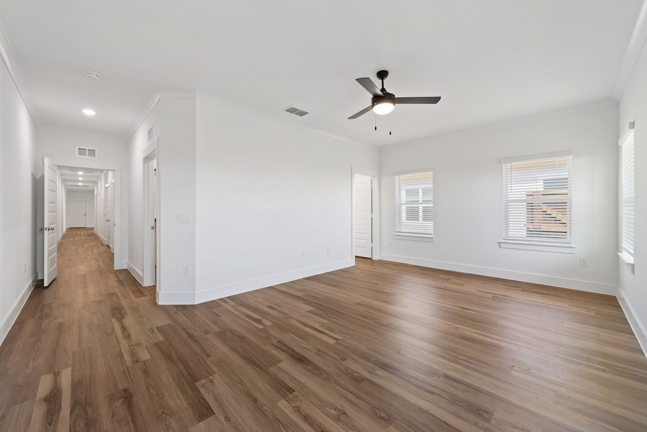 Modern living space featuring sleek hardwood floors and abundant natural light from large windows, with a view of a long hallway and ceiling fan.