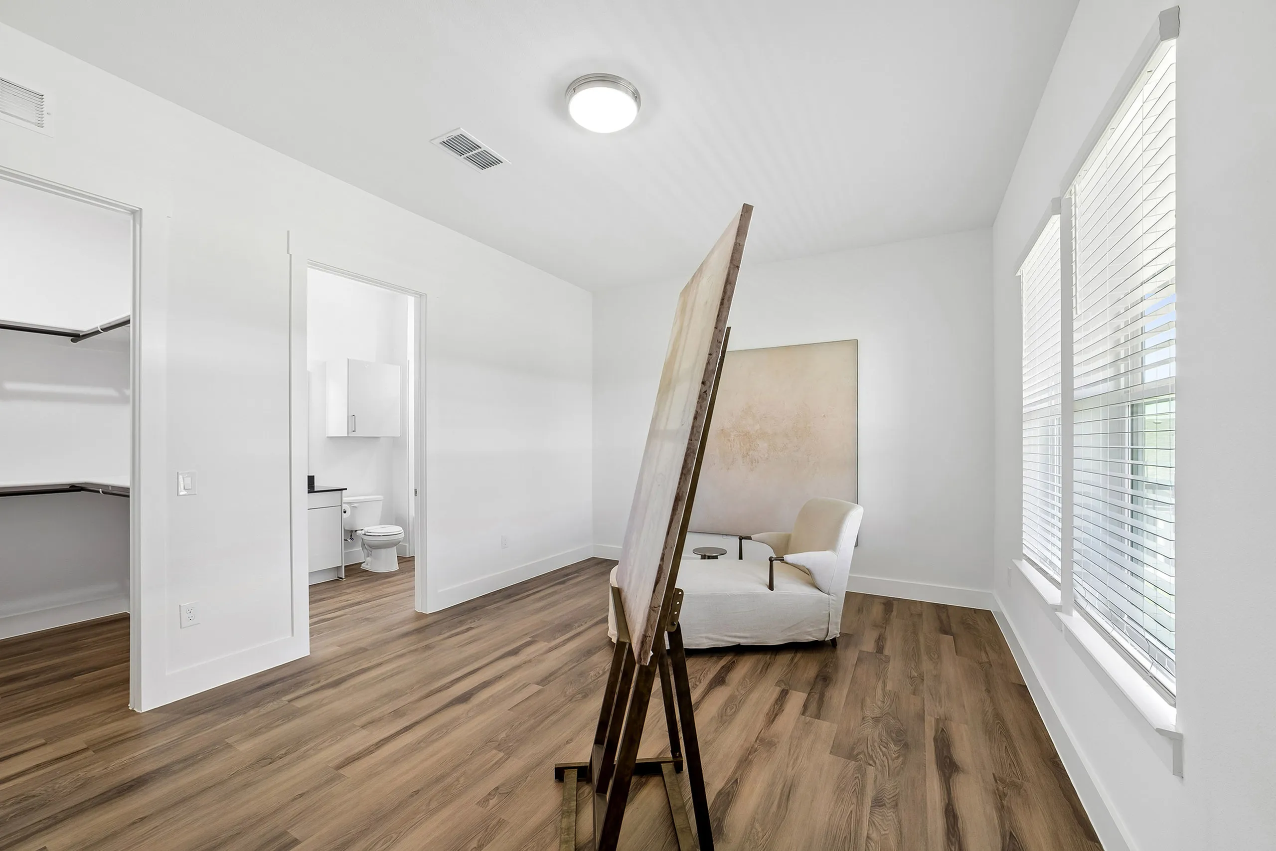 Bright, modern room with wooden flooring showcasing a cozy corner with an armchair, canvas stand, and ample natural light through wide windows. Adjacent walk-in closet and sleek bathroom visible.