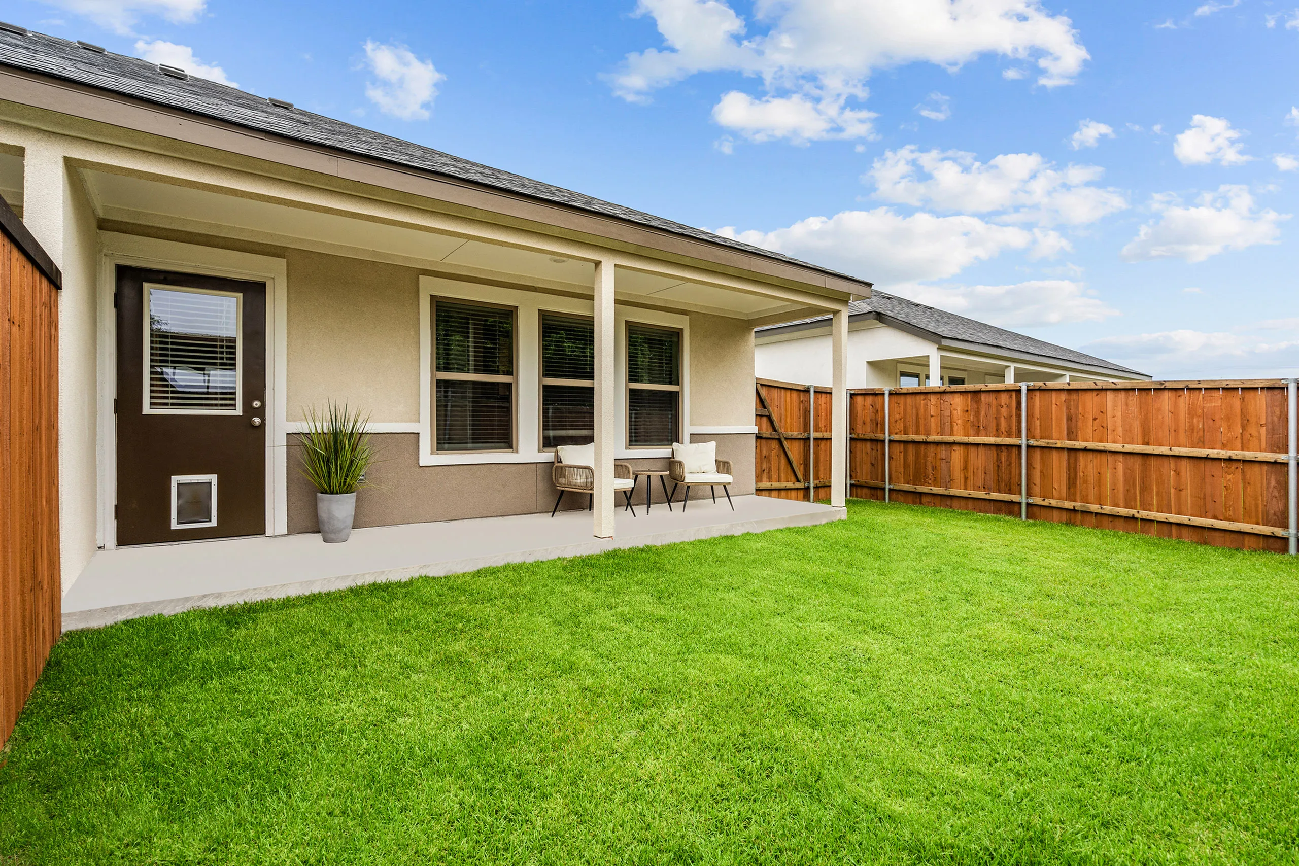 Charming backyard of a modern home featuring a cozy covered patio with seating, lush green lawn, and wood fence under a bright blue sky.