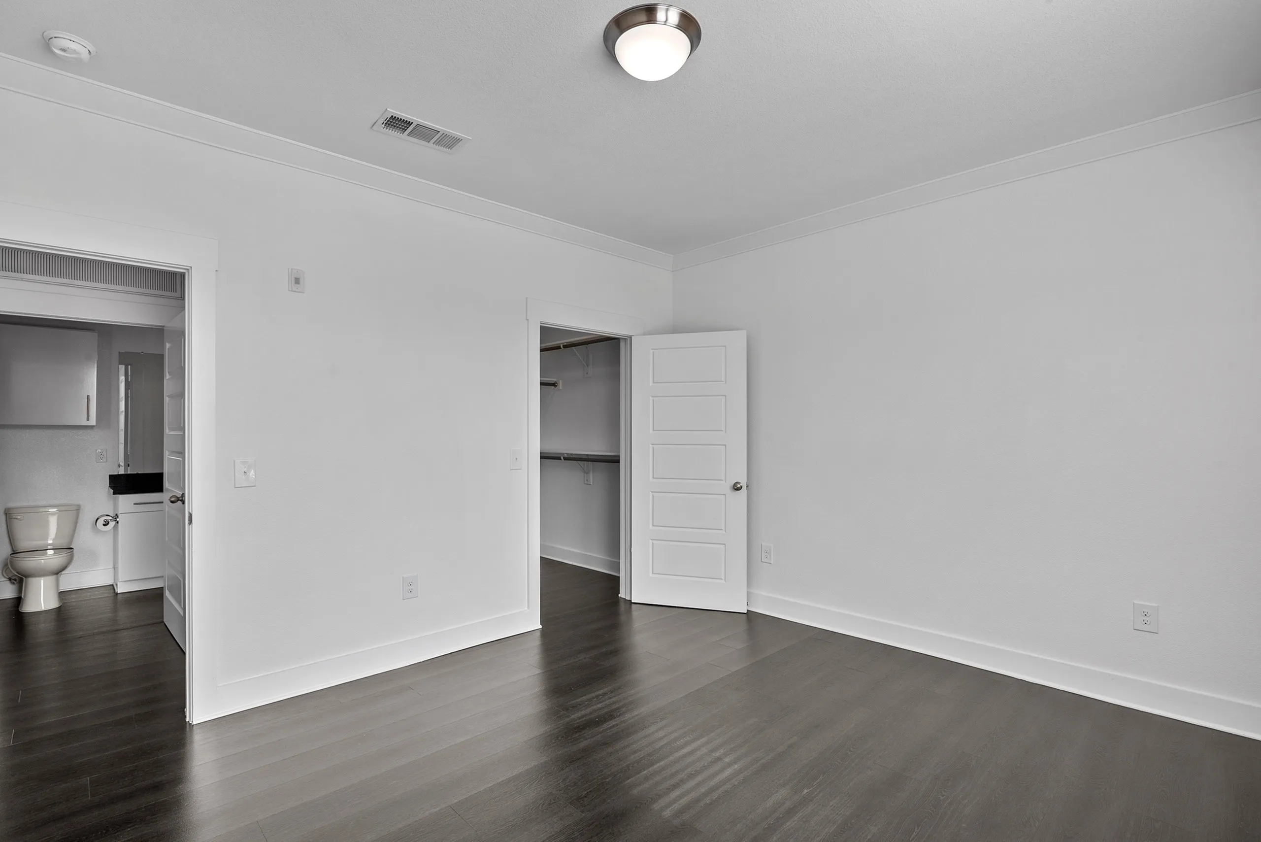 Spacious empty room with sleek hardwood floors, a modern ceiling light, and access to a bathroom and walk-in closet.