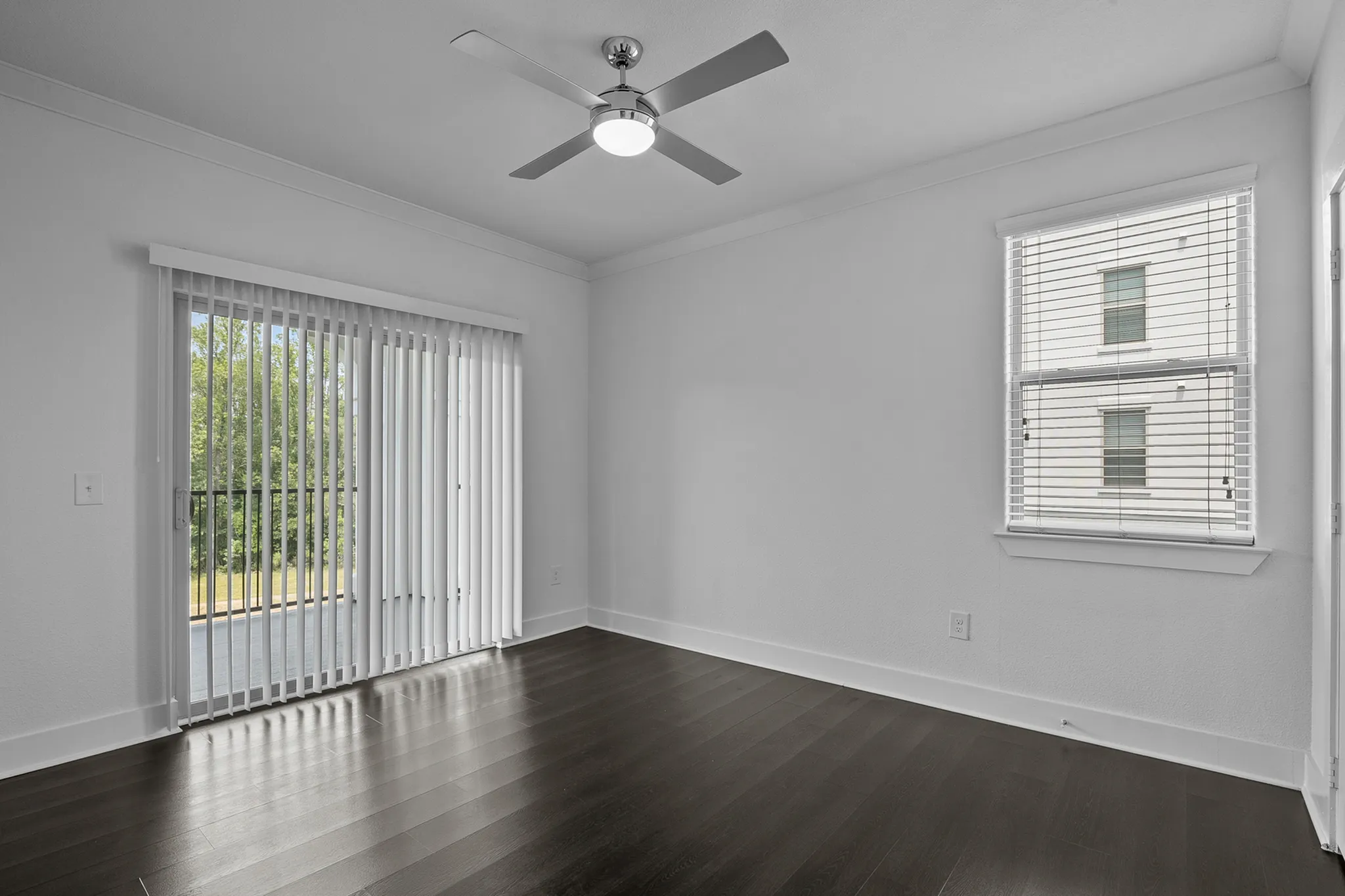 Bright room with sleek dark hardwood floors, modern ceiling fan, and large windows with vertical blinds opening to a serene balcony view.