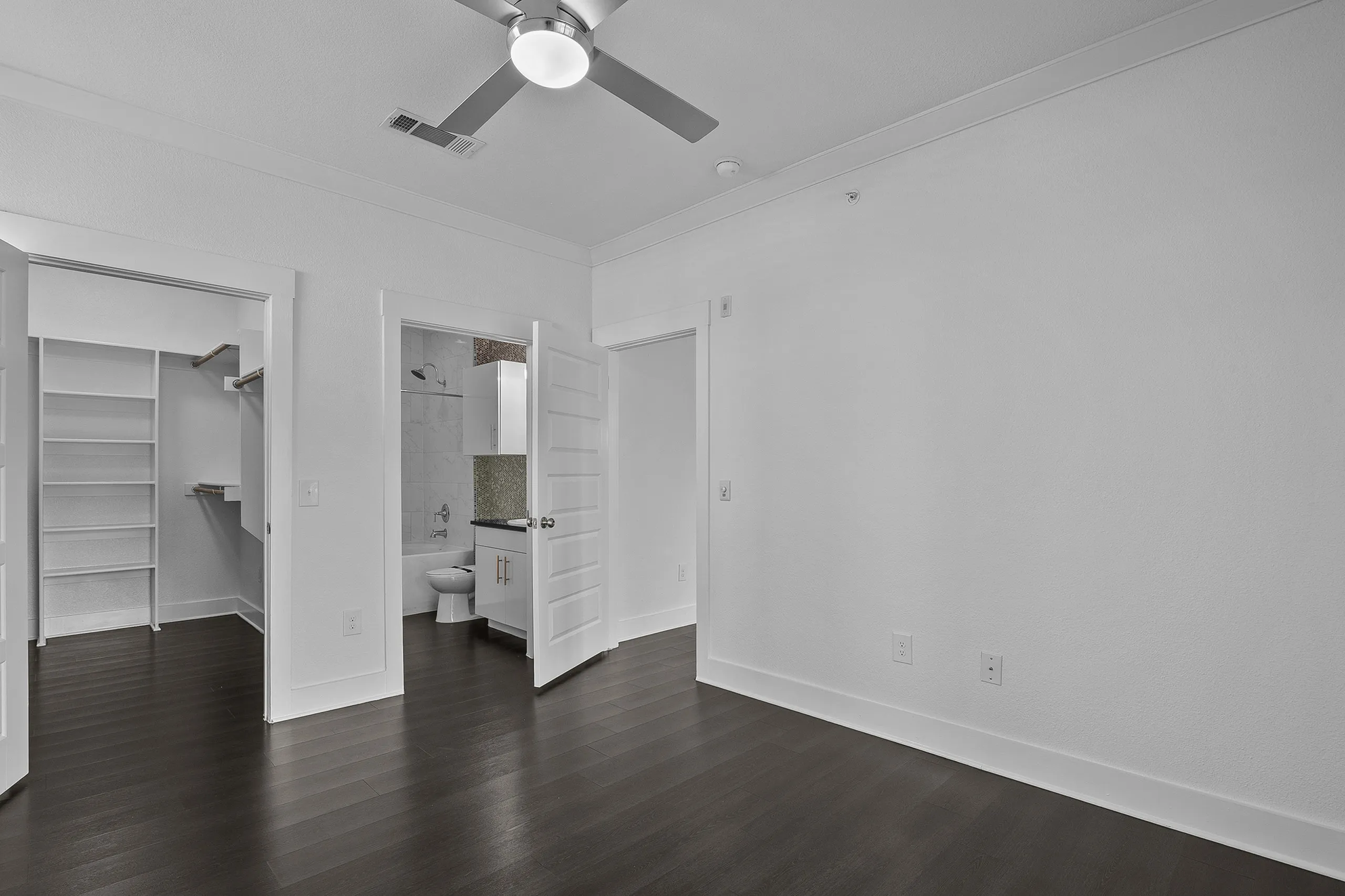 Modern bedroom with sleek ceiling fan and direct access to walk-in closet and bathroom, featuring elegant dark hardwood floors.