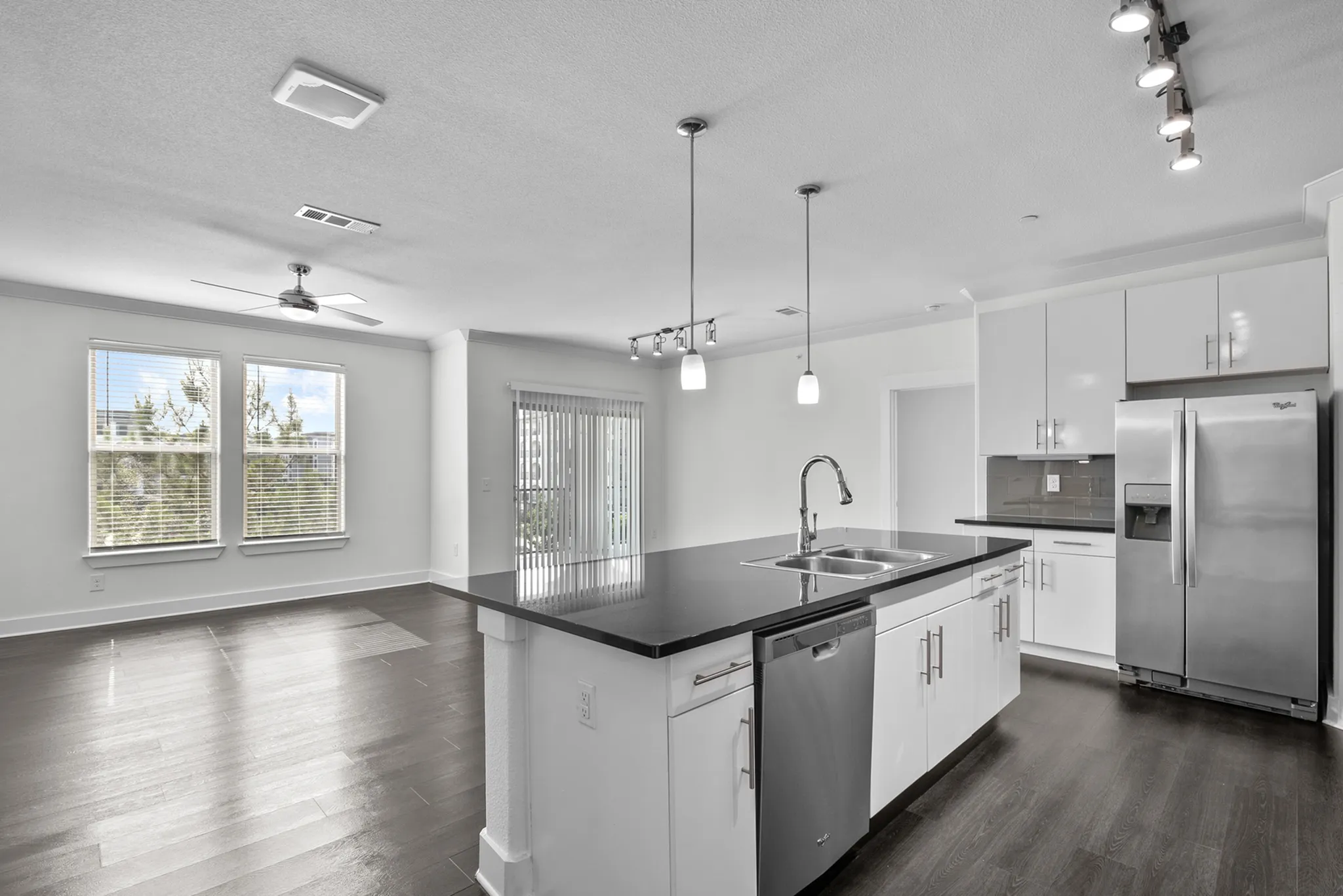 Modern open-concept apartment kitchen with sleek stainless steel appliances and elegant countertops, leading into a spacious, light-filled living area with wood flooring and large windows.