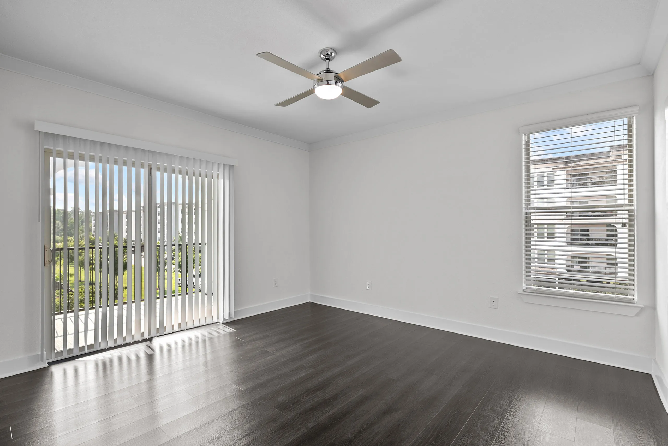Bright apartment room with sleek dark wood flooring, large glass sliding doors to a balcony, and modern ceiling fan, offering a serene view of green surroundings.