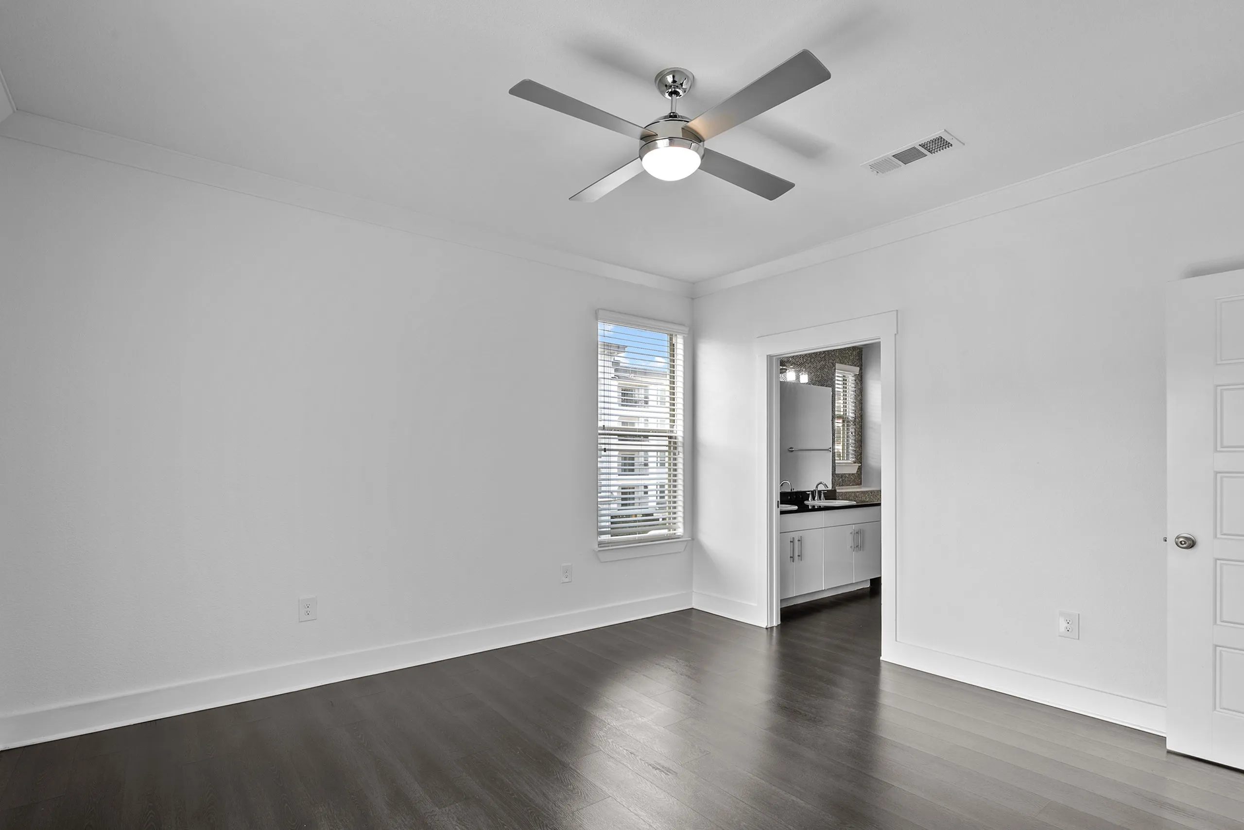 Bright, modern room with sleek dark wooden flooring, large window for natural light, and ceiling fan; leading to a stylish kitchenette with white cabinets.