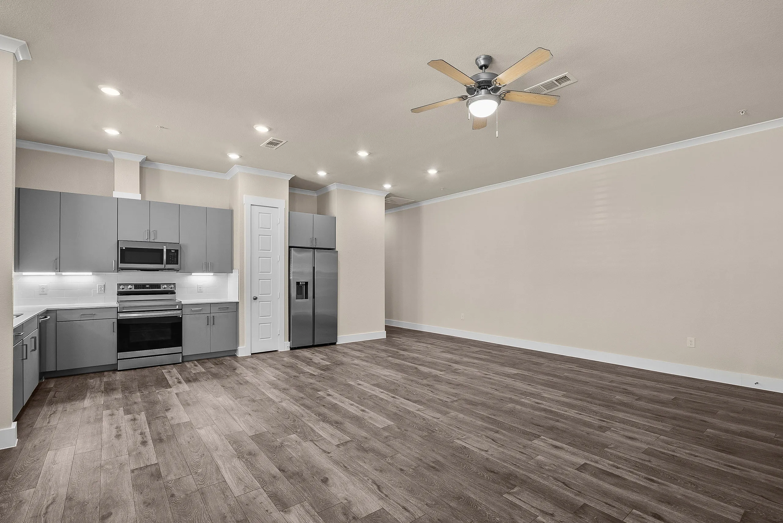 Modern open-concept kitchen with sleek gray cabinets, stainless steel appliances, and elegant wood flooring, flowing into a spacious, well-lit living area with a ceiling fan.