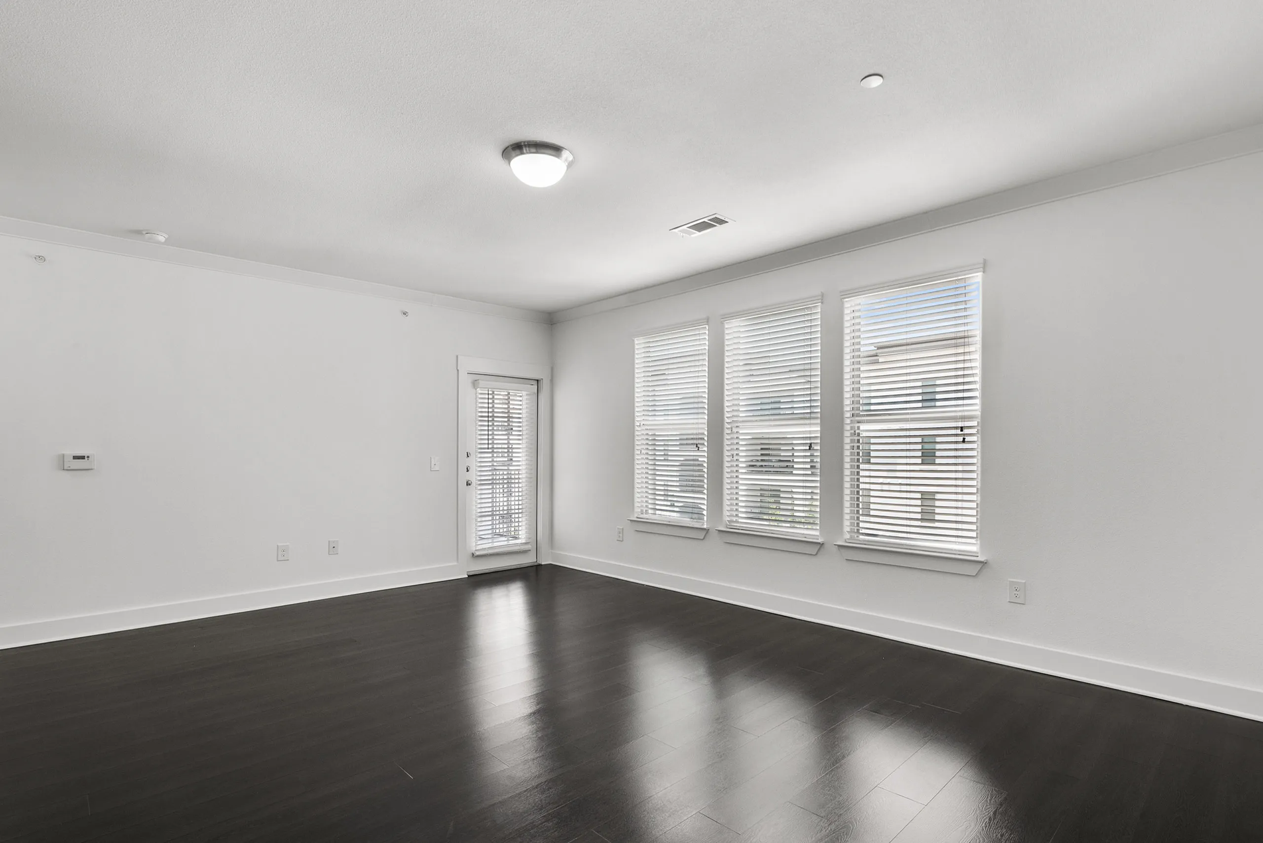 Bright and spacious living room featuring large windows with blinds and sleek dark wood flooring.
