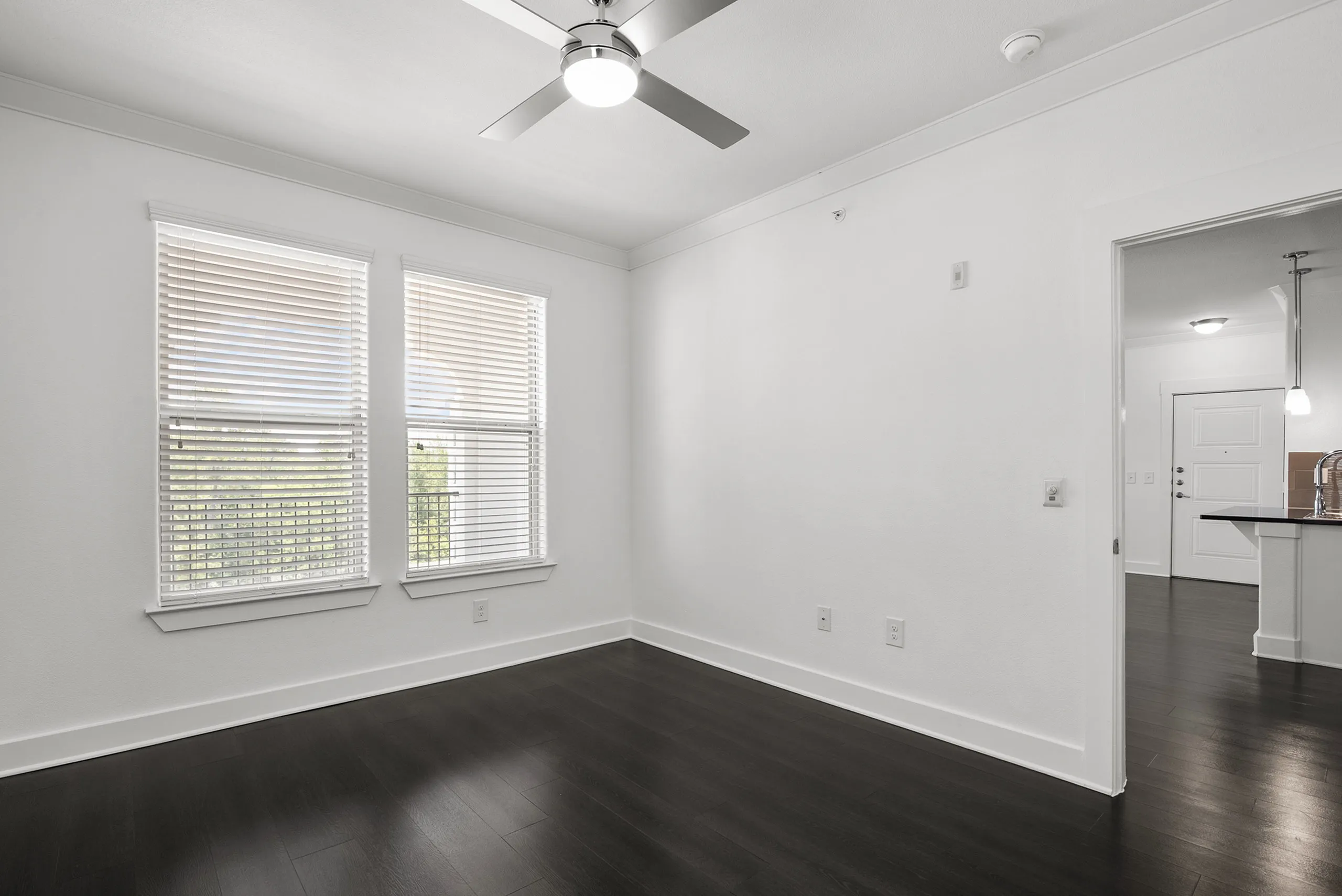 Bright room with sleek dark wood flooring, large windows with blinds, and modern ceiling fan, adjacent to a stylish kitchen area.