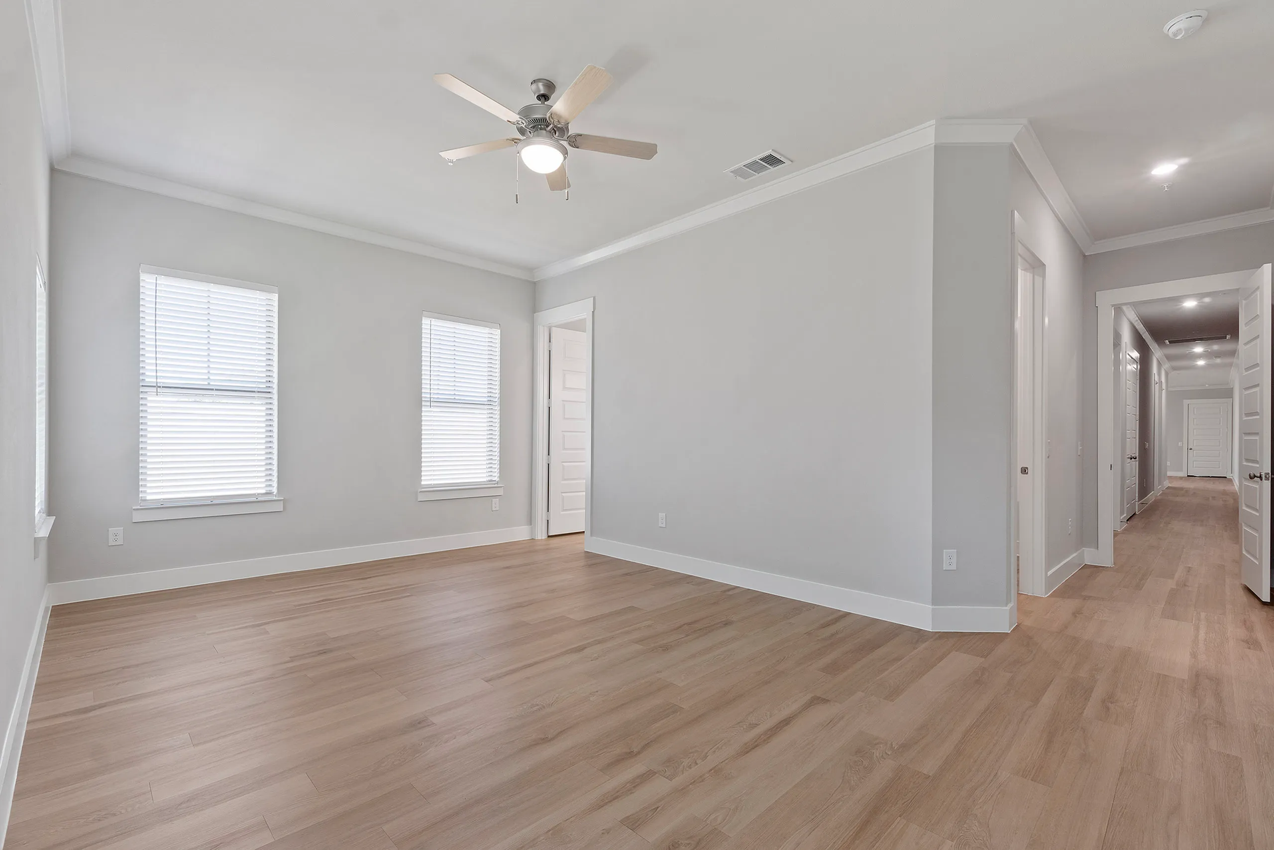 Bright and spacious empty room with elegant wood flooring, modern ceiling fan, and ample natural light from large windows, perfect for a personalized living space.
