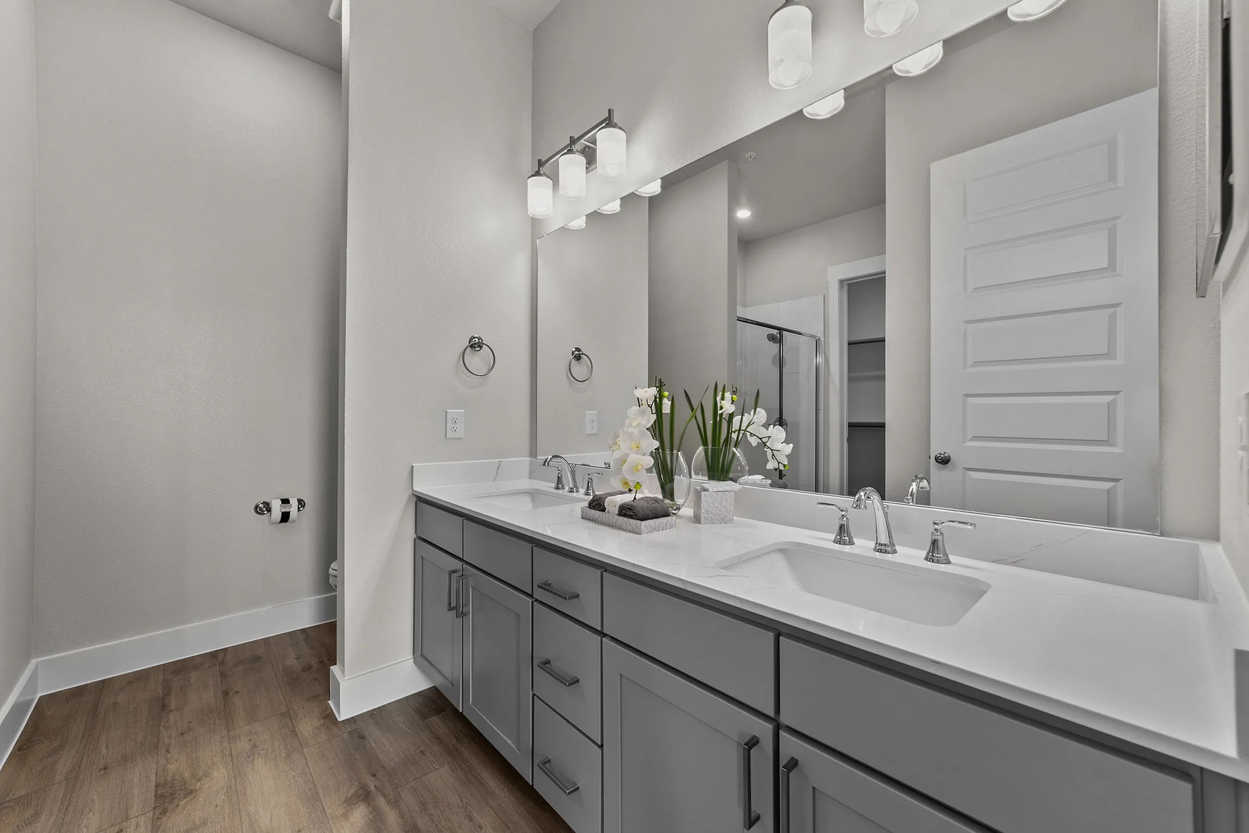 Elegant modern bathroom featuring double sinks, sleek cabinetry, and stylish lighting fixtures with a large mirror and wood flooring.