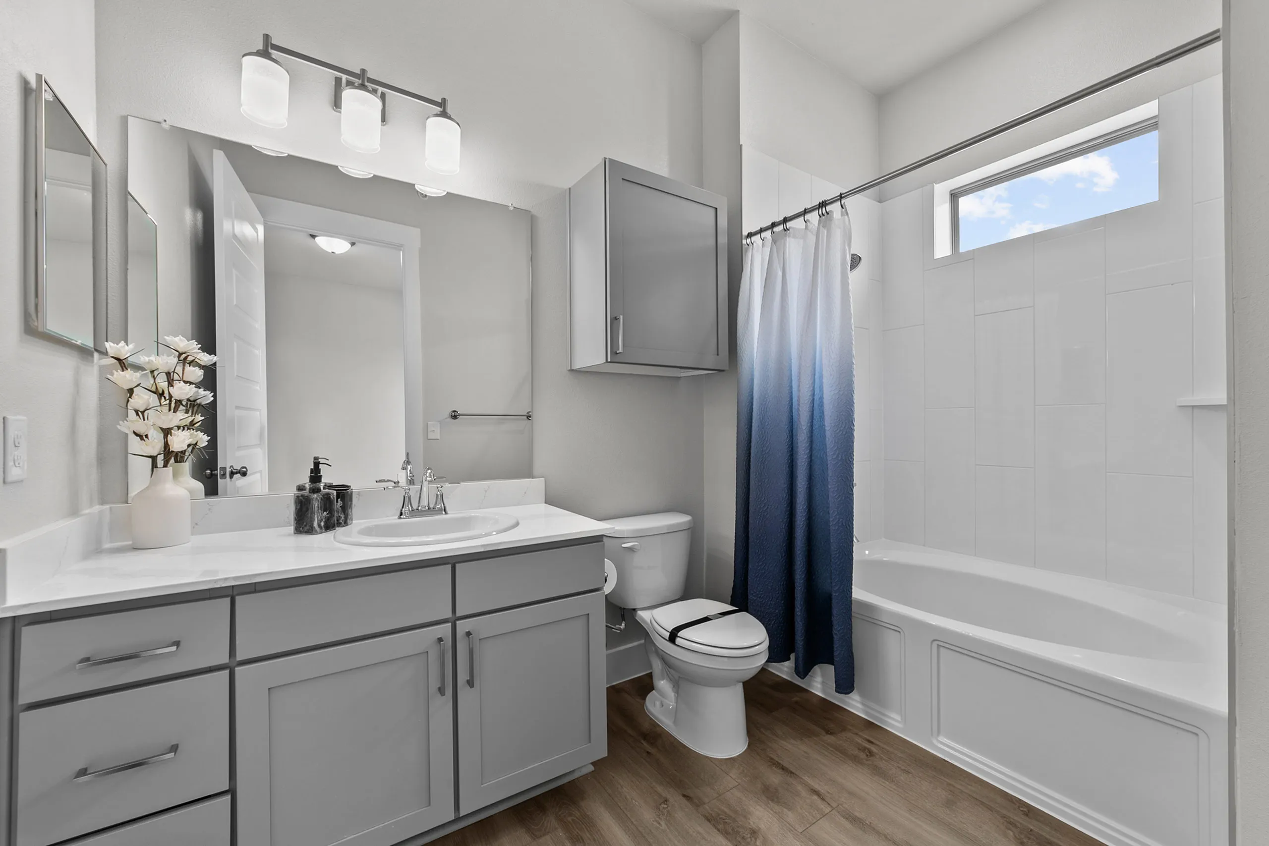 Modern bathroom with sleek grey cabinetry, elegant fixtures, and a stylish gradient shower curtain, enhanced by natural light from a high window.