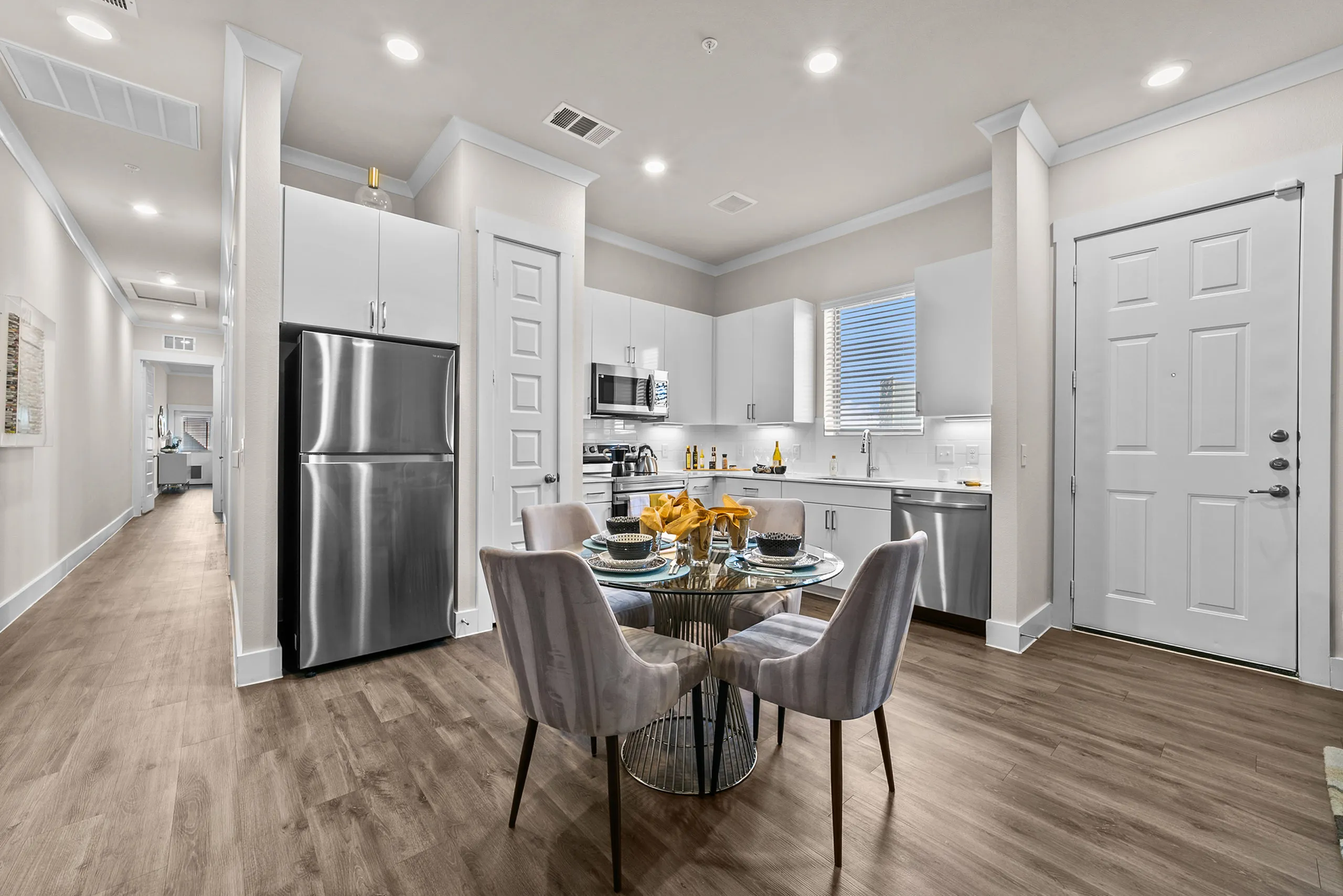 Modern kitchen and dining area with sleek white cabinets, stainless steel appliances, and an elegant glass table set for four.
