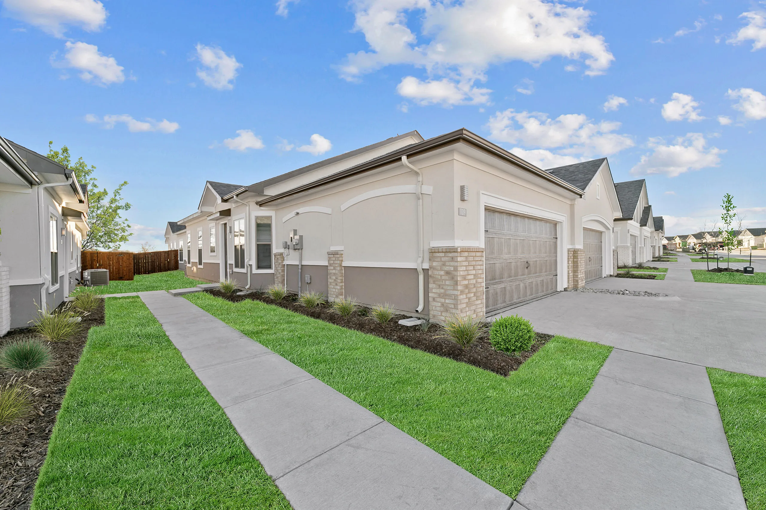 Charming suburban street view with modern single-story homes featuring brick accents, pristine lawns, and a clear, blue sky.
