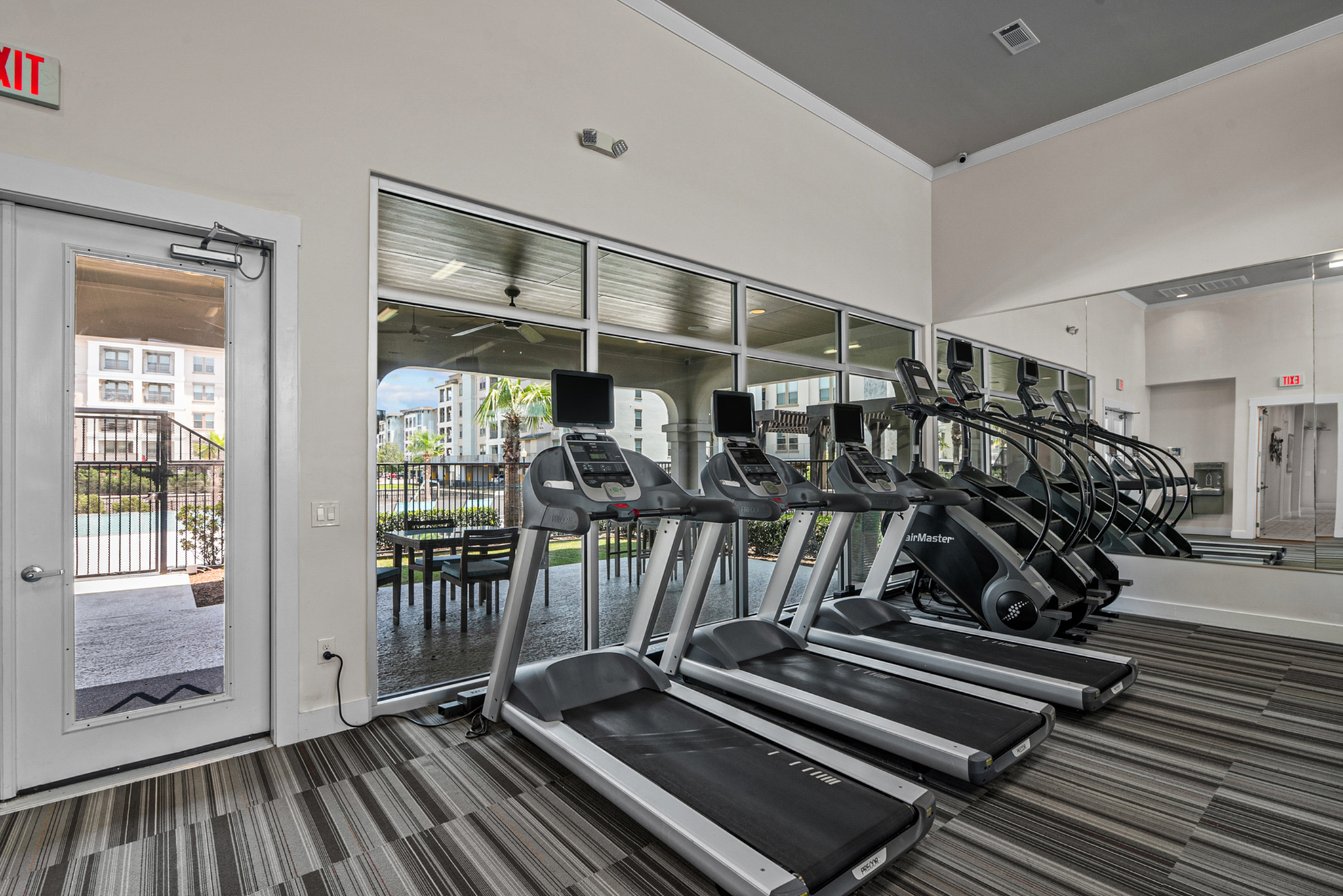 Modern fitness center with a row of sleek treadmills, mirrored wall, and natural light streaming through large windows overlooking a vibrant outdoor patio.