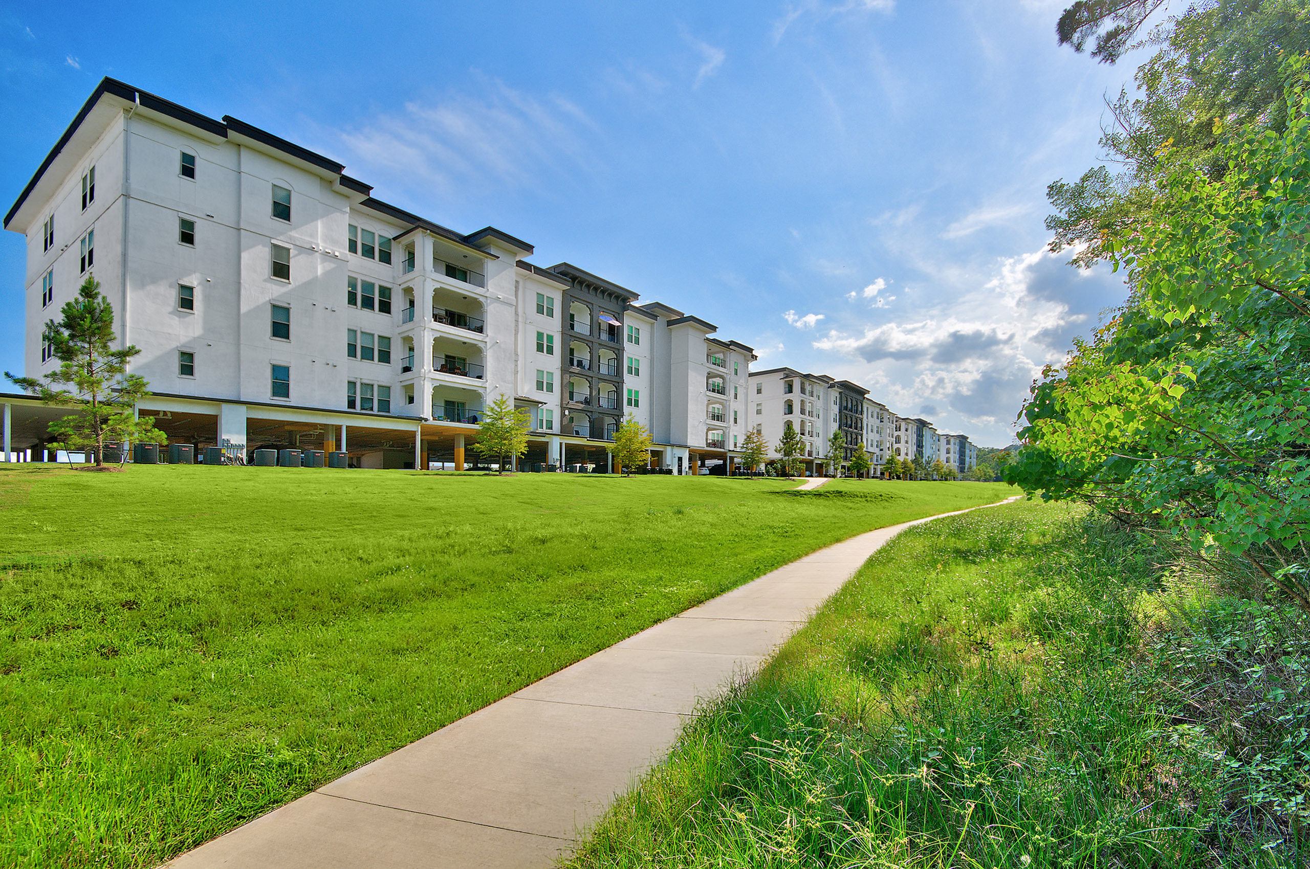 Modern apartment complex with elegant architecture, expansive green lawn, and a scenic walkway under a clear blue sky.