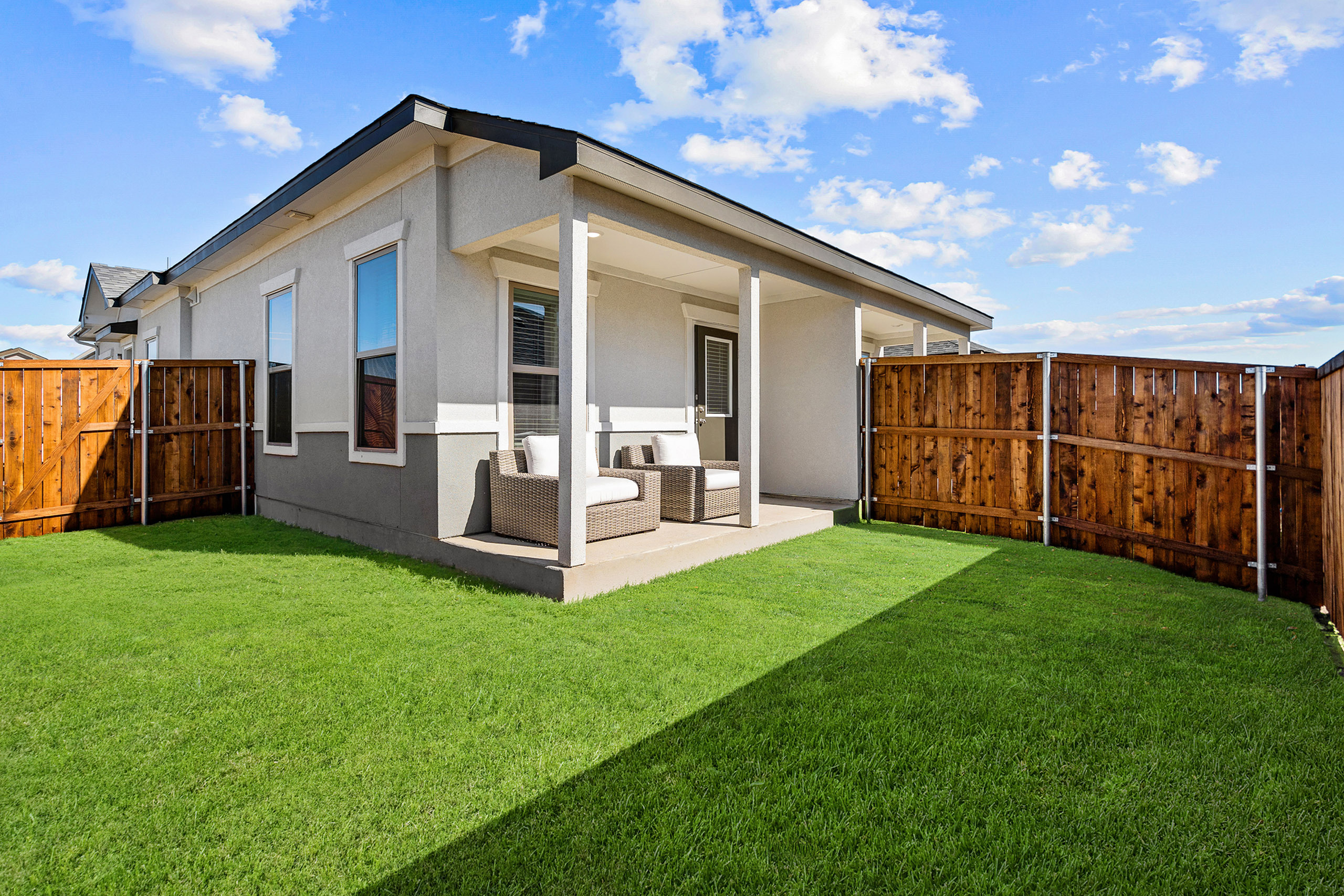 Charming modern home with a cozy patio, lush green lawn, and wooden privacy fence under a bright blue sky.