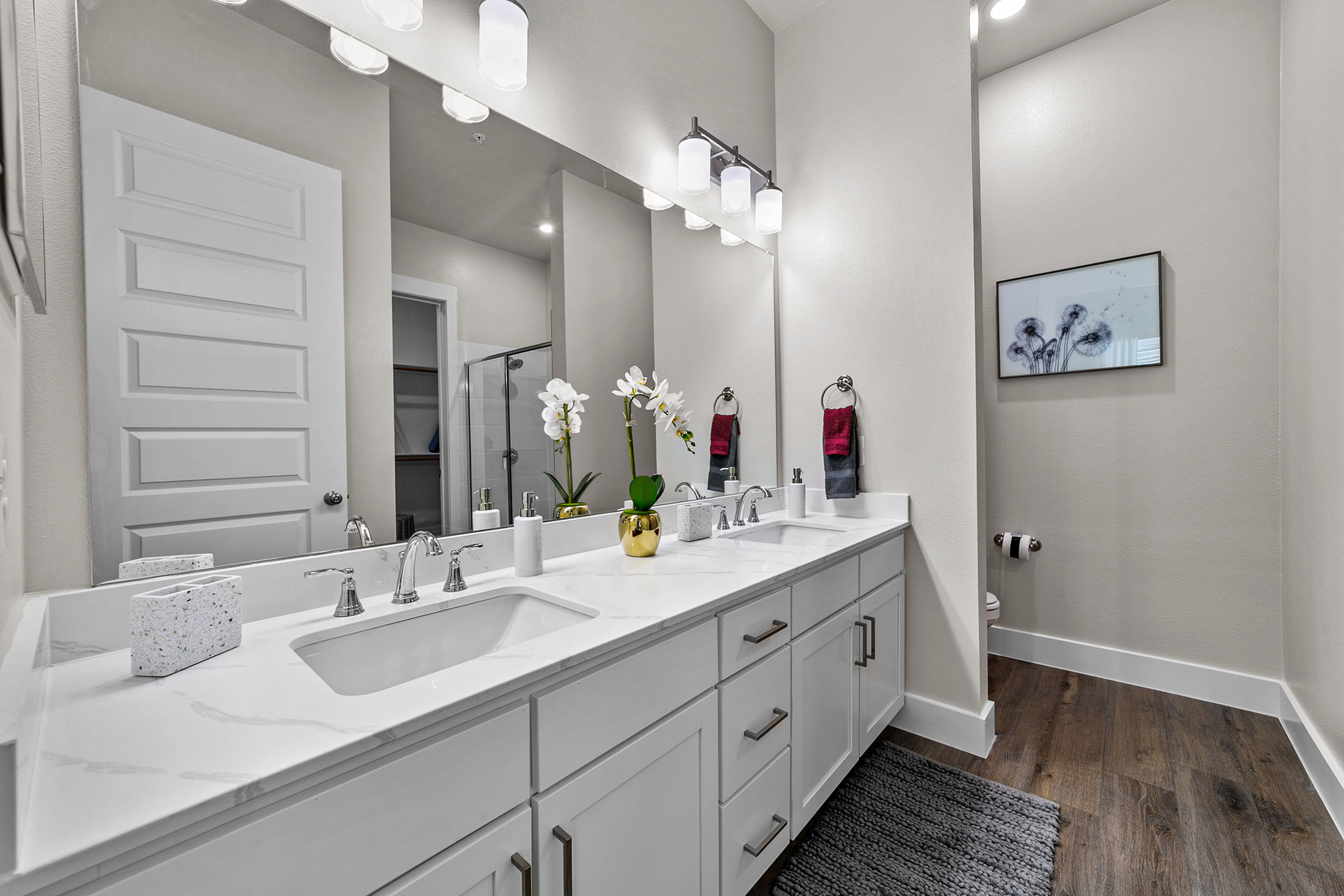 Elegant bathroom featuring double sinks, sleek white cabinetry, a spacious mirror, and stylish lighting, complemented by a modern glass shower and wood flooring.