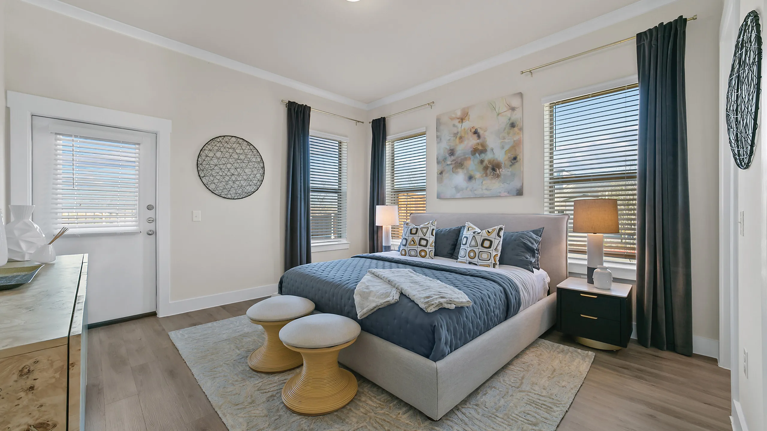 Elegant bedroom with chic decor, featuring a plush blue bedspread, modern art, and abundant natural light from multiple windows.