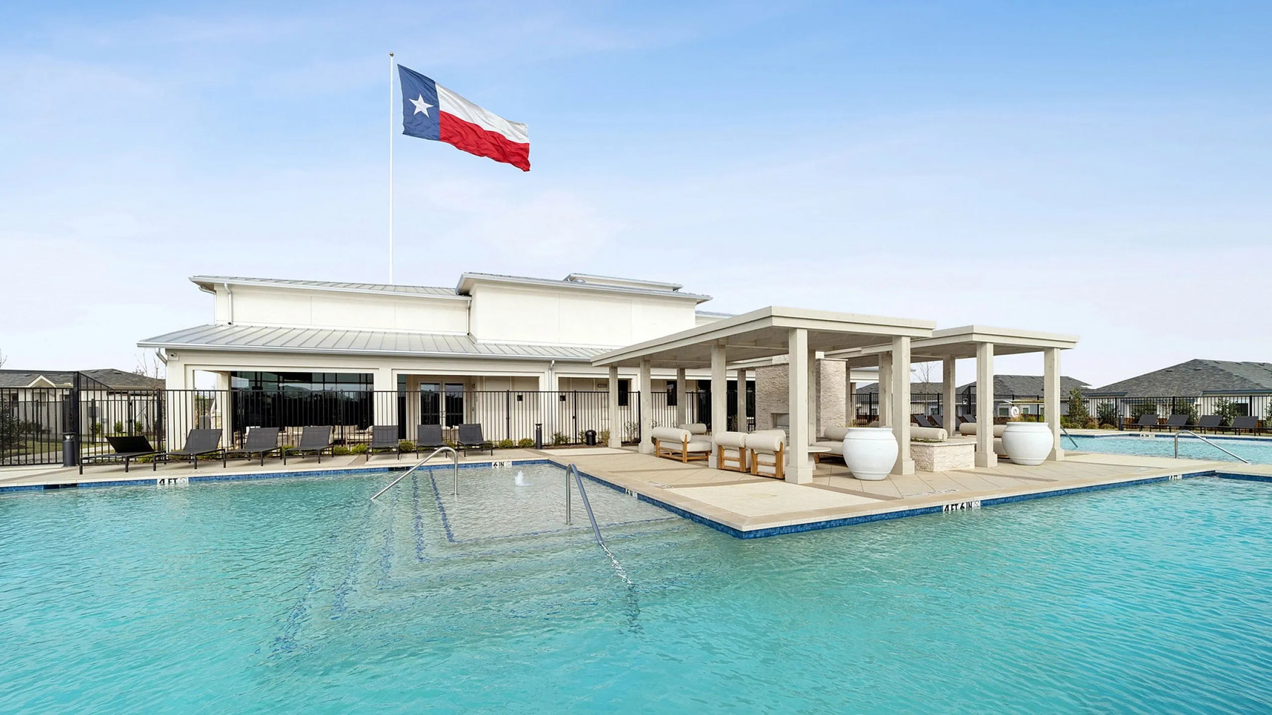 Luxurious apartment community pool area with modern cabanas and expansive lounging space, graced by a large state flag in the background.