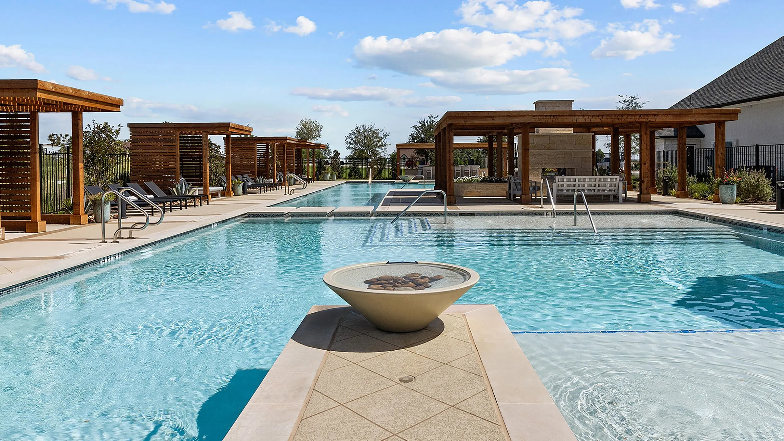 Luxurious outdoor pool area featuring elegant wooden cabanas, pristine water, and a modern fire pit, set against a vibrant blue sky.
