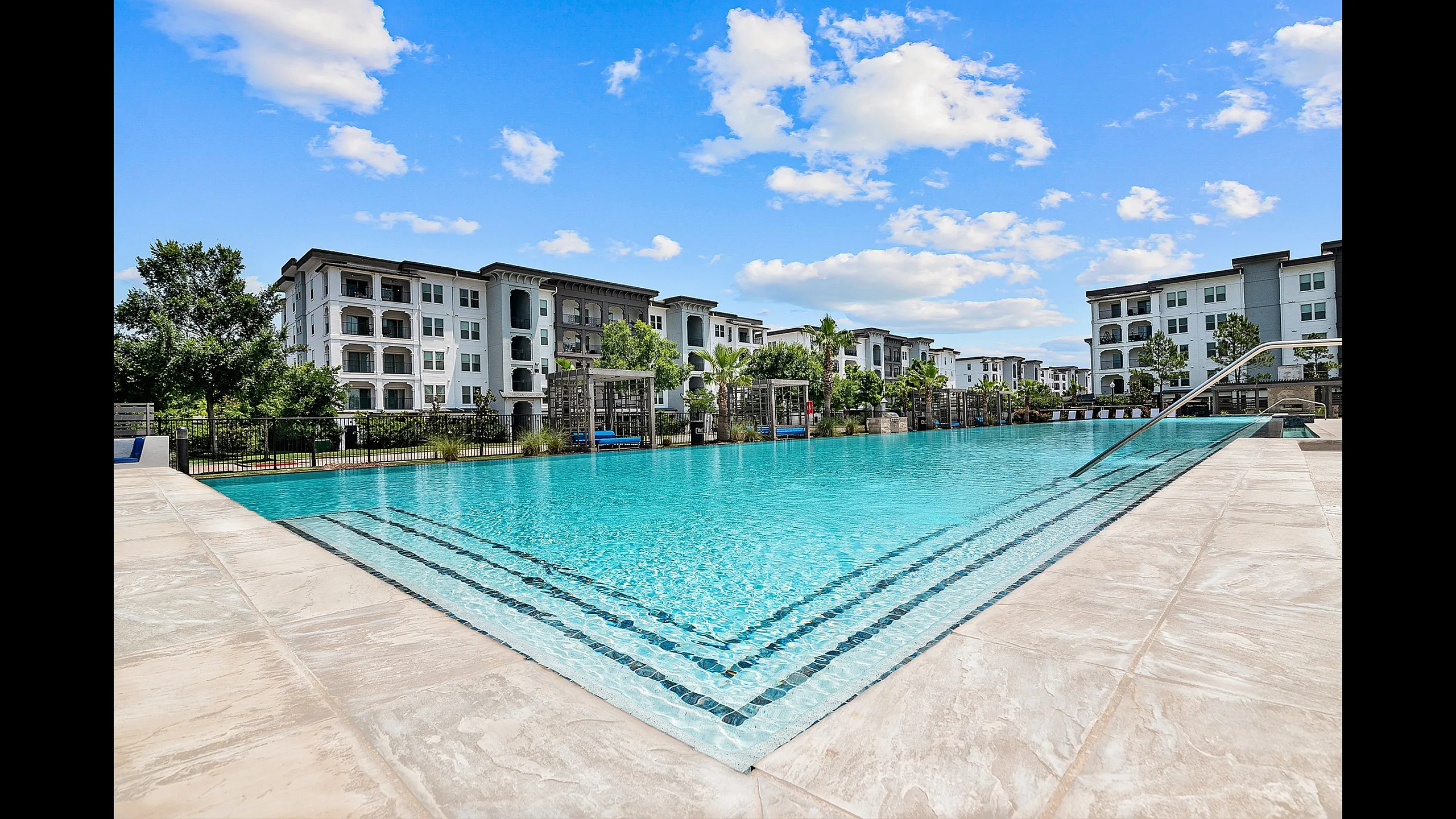 Luxury apartment complex with a stunning outdoor pool, surrounded by modern architecture and lush greenery under a bright blue sky.