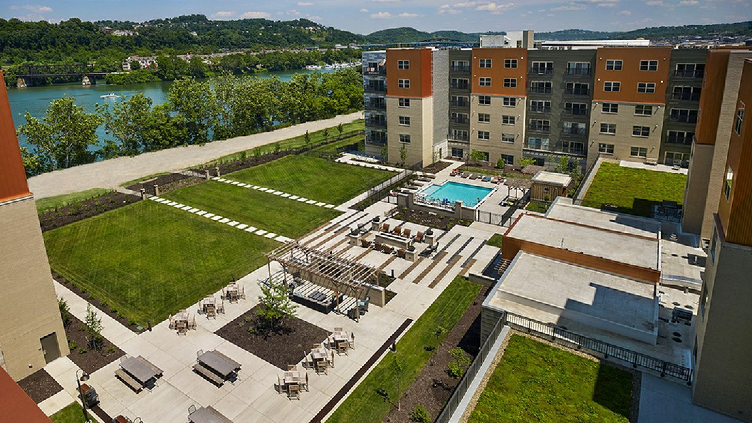 Luxurious apartment complex with a beautiful courtyard, lush greenery, outdoor seating, and a pristine swimming pool overlooking a scenic river view.