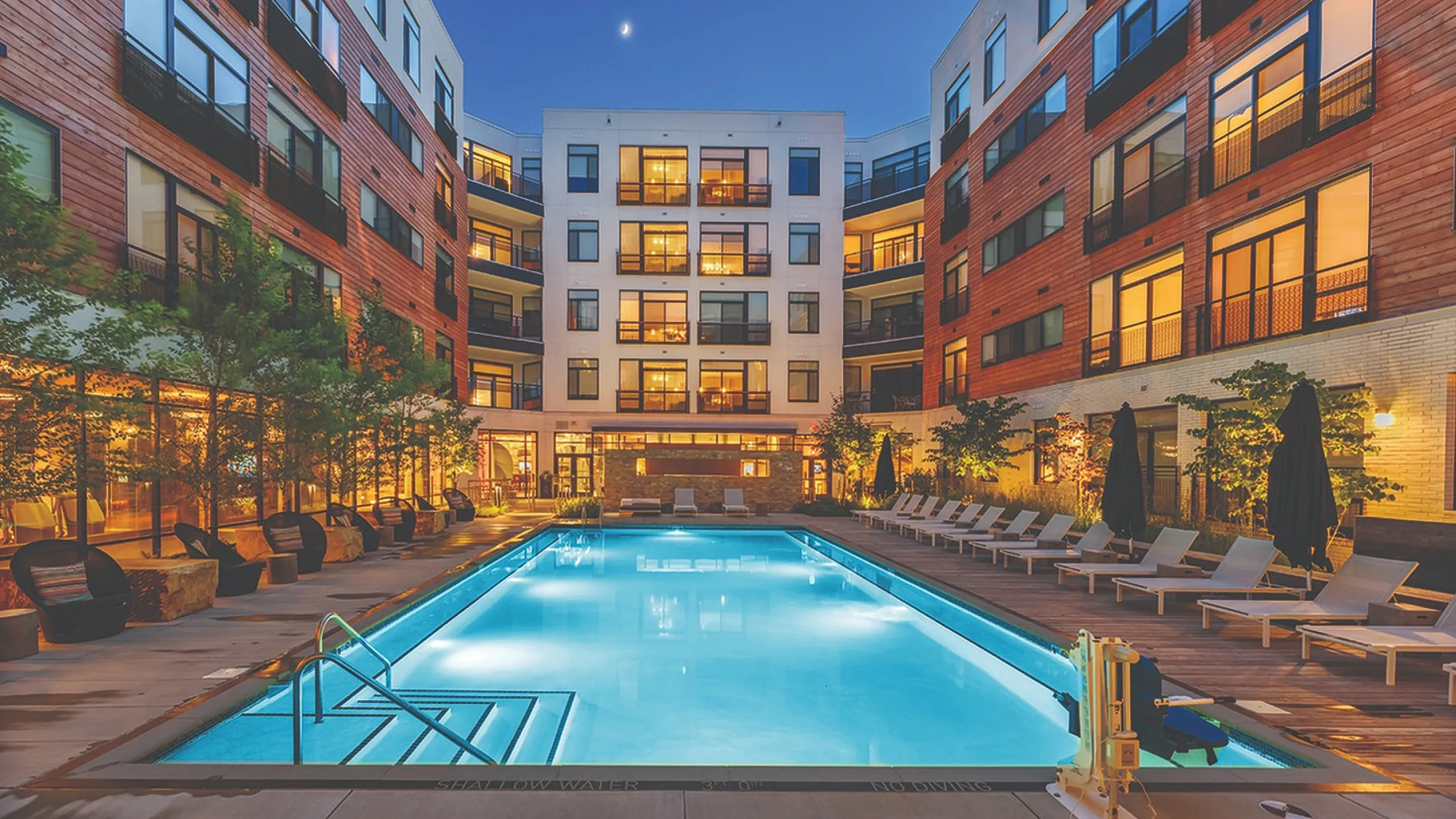 Luxurious apartment complex featuring a sparkling illuminated pool, surrounded by modern architecture and lush greenery, perfect for relaxation and enjoyment under the night sky.