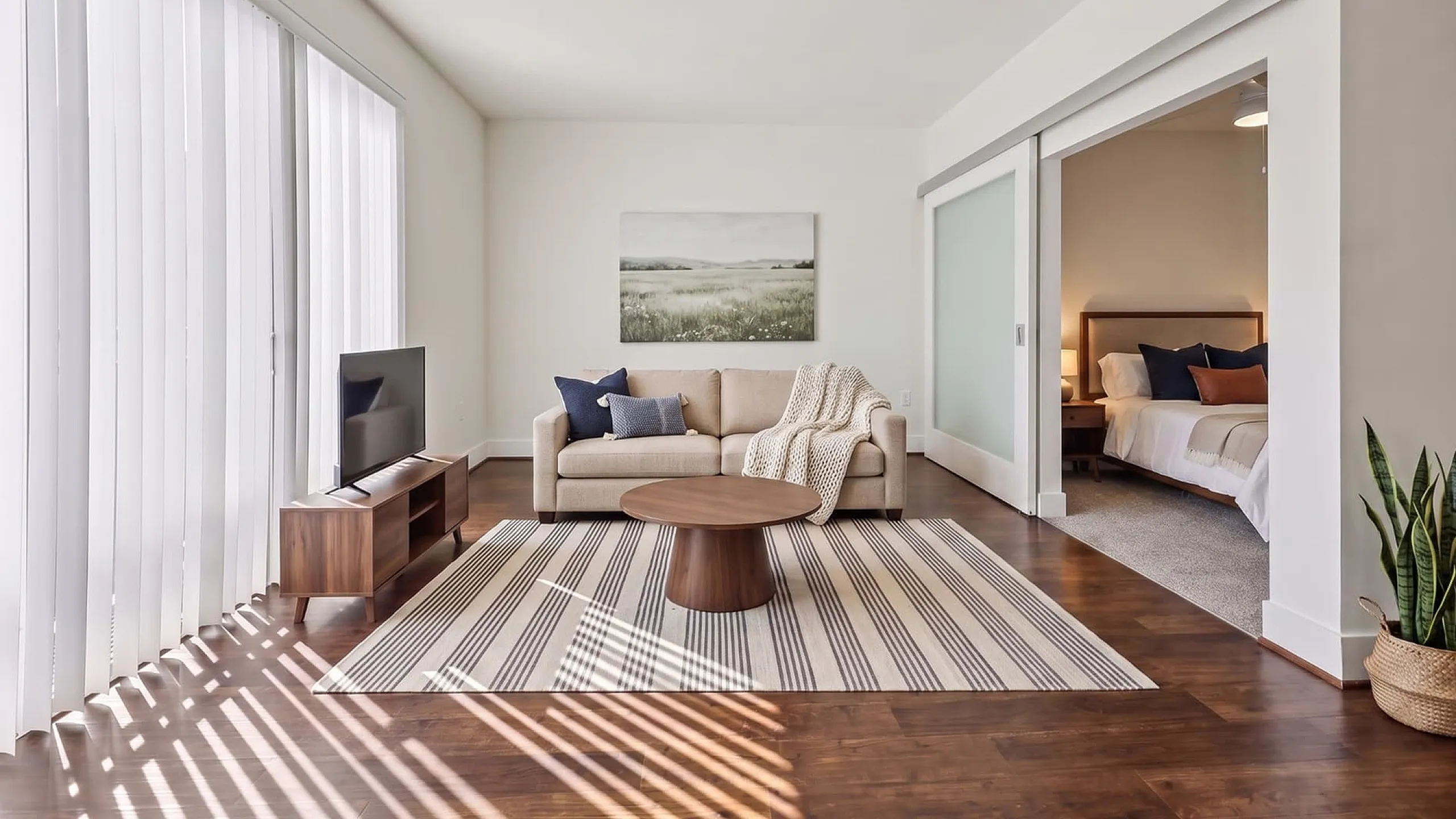 Bright and airy living area with stylish decor, featuring a comfortable beige sofa, modern coffee table, and hardwood floors leading to a cozy bedroom.