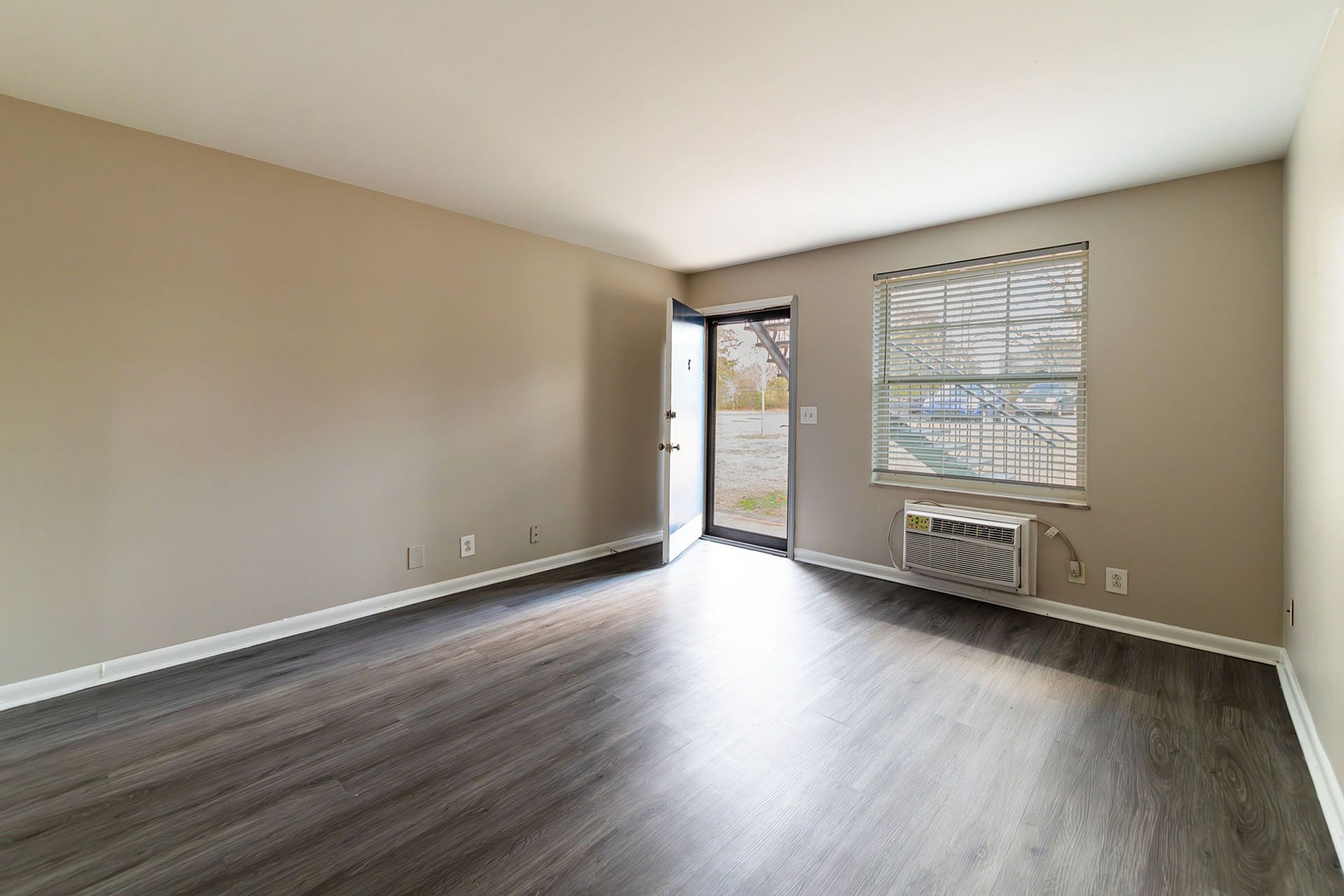 Bright and airy empty living room with wood flooring, large window, and front door access.