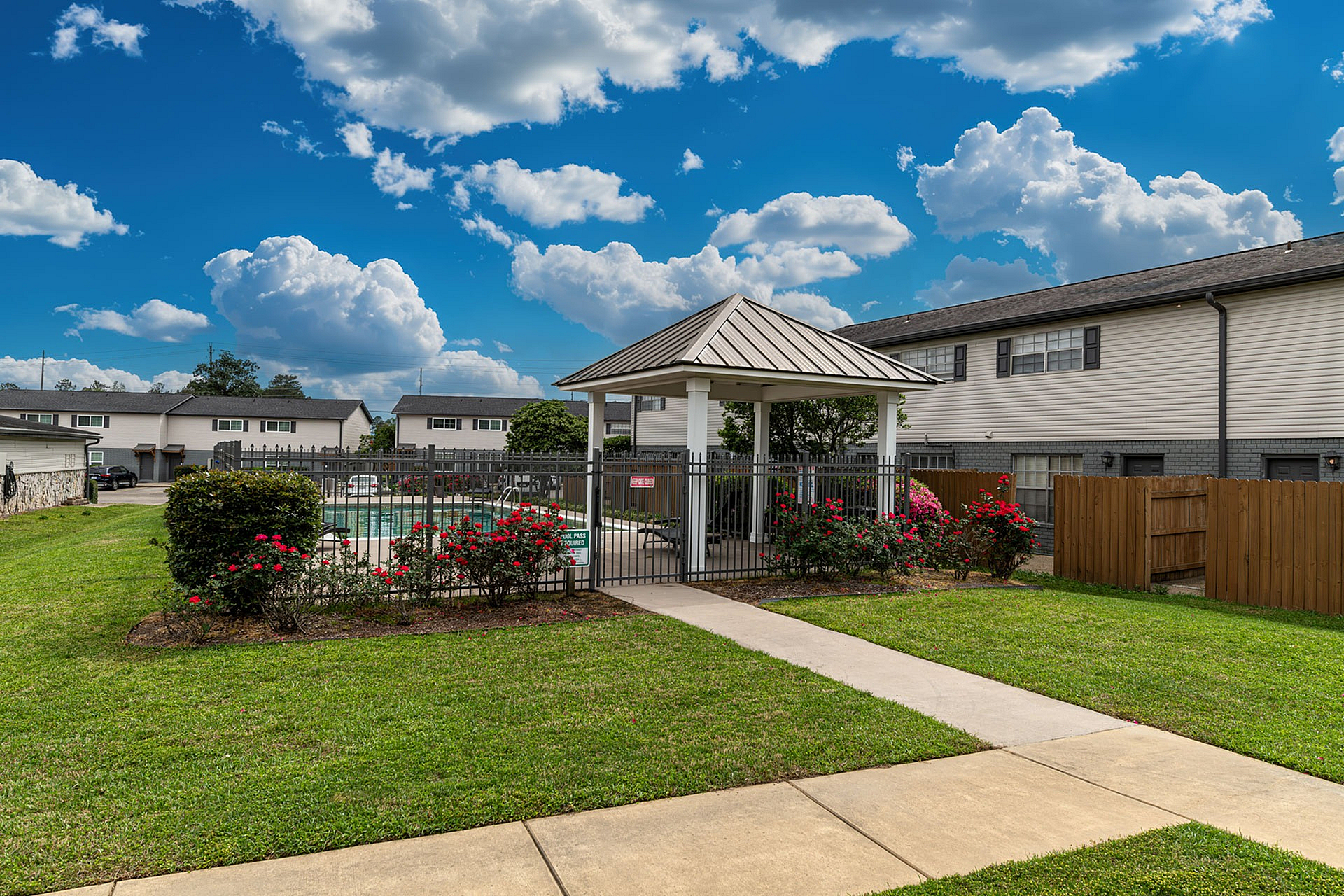 Charming pool area with a covered pavilion, surrounded by vibrant roses and lush greenery, set against a backdrop of modern townhouses and a striking blue sky with fluffy clouds.