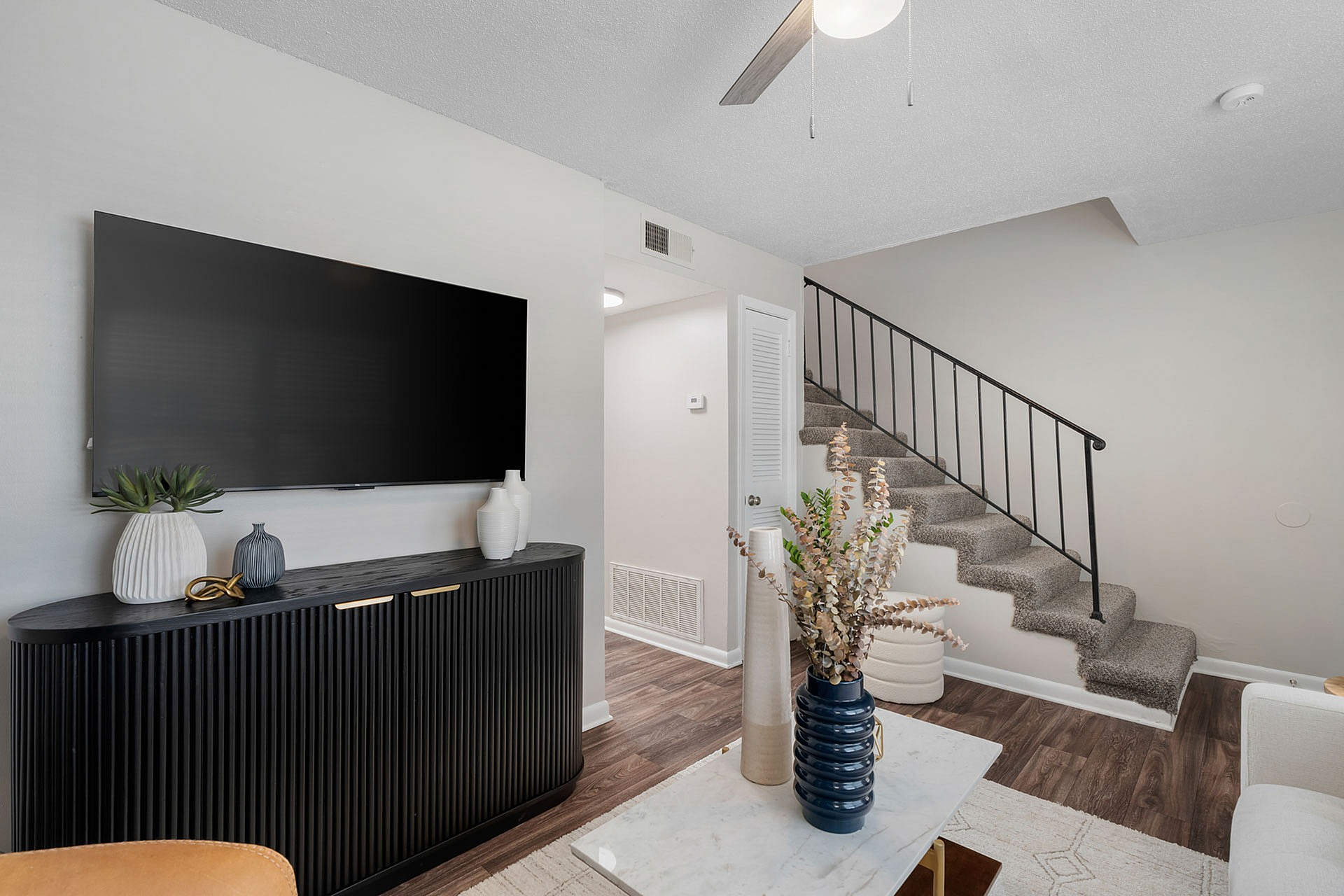 Chic modern living room with a sleek black entertainment console, large flat-screen TV, and elegant staircase accentuated by tasteful decor and vibrant greenery.