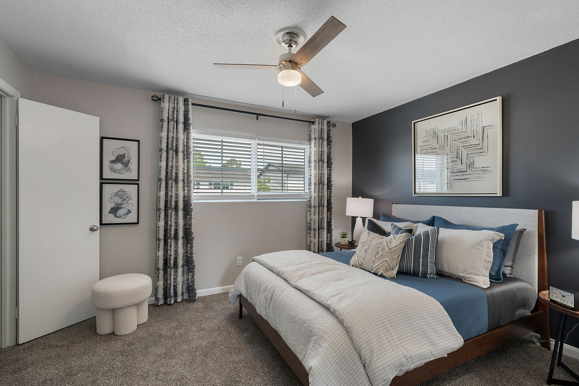 Elegant bedroom with modern decor, featuring a cozy bed with blue and beige linens, stylish wall art, and natural light through large windows.