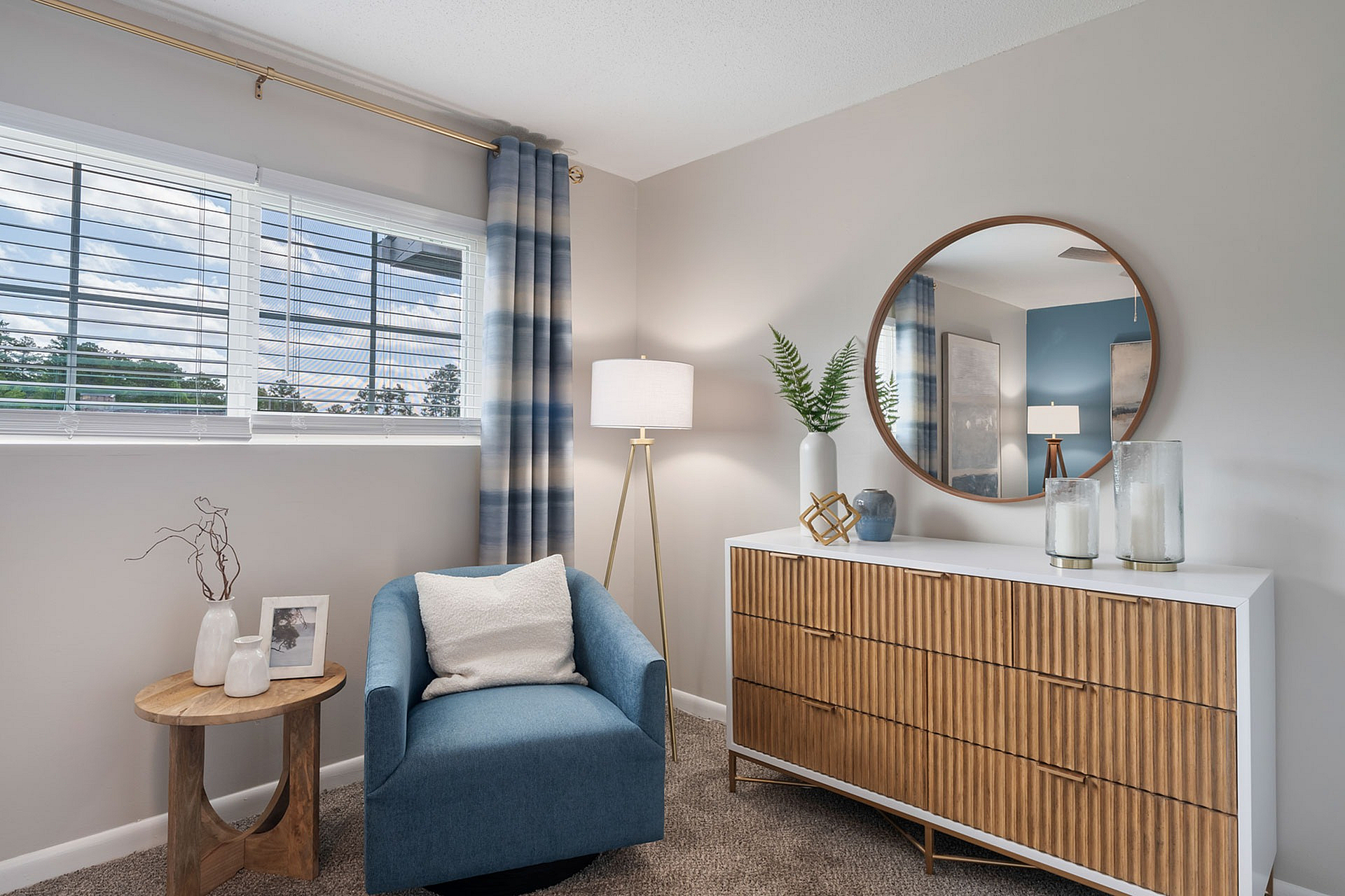 Chic living space featuring a cozy blue armchair, elegant wooden dresser with a round mirror, and stylish decor under soft natural light.