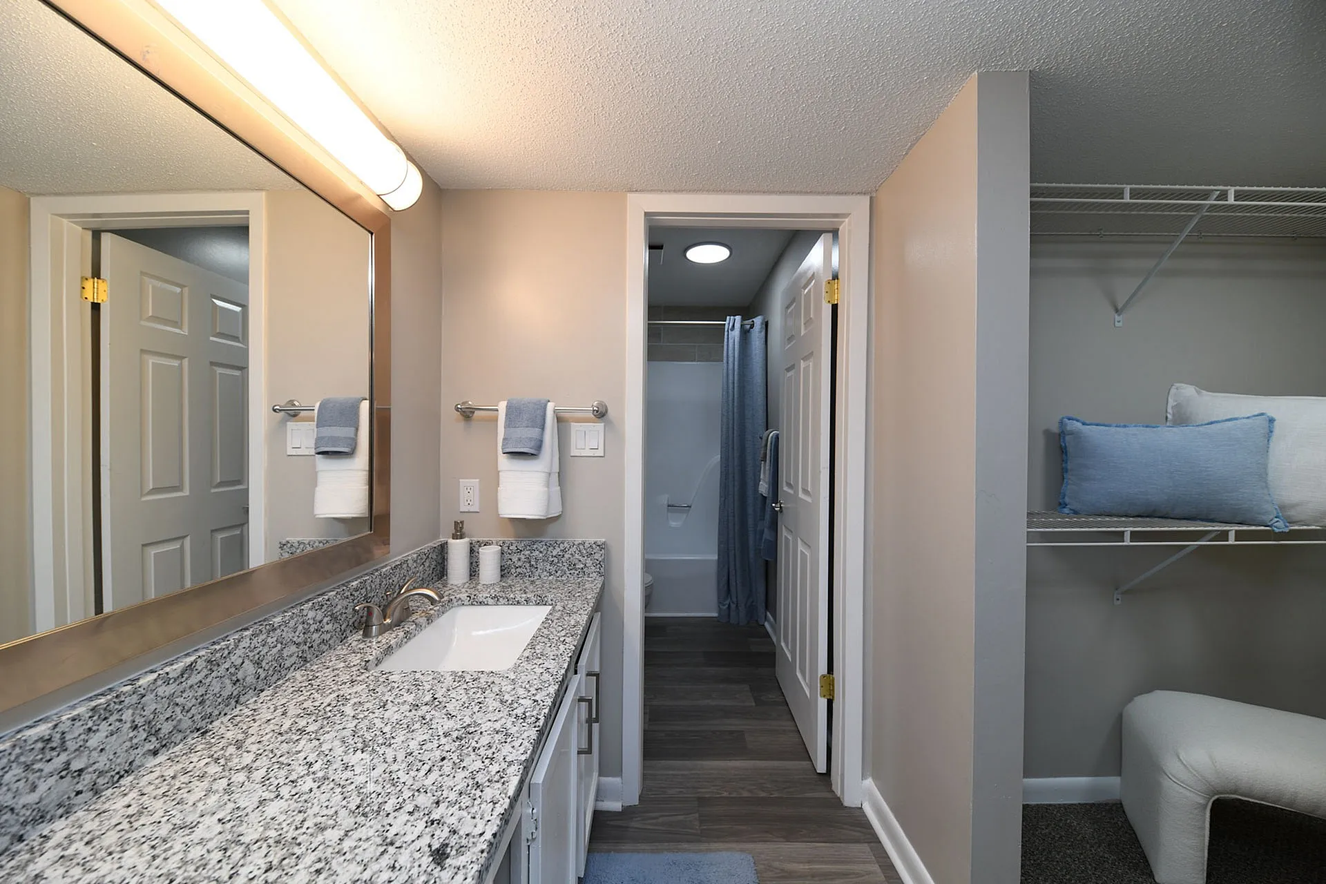 Elegant bathroom with a granite countertop, large mirror, and towel racks, leading to a modern shower area and adjacent closet space with stylish shelving.